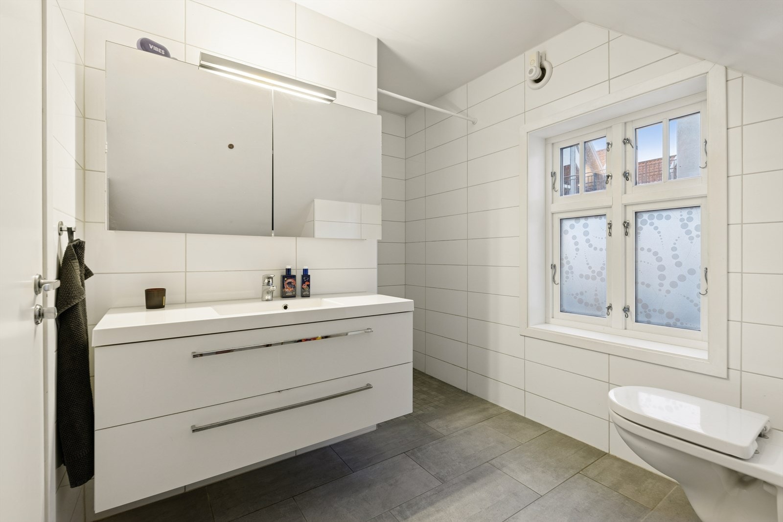 Rommet er innredet med en baderomsinnredning med nedfelt servant, en dusjnisje og et vegghengt toalett. Ventilasjonen består av en elektrisk vifte i veggen og en luftespalte over døren. Boligens varmtvannstank på ca. 120 liter er plassert på badet. Galleribilde