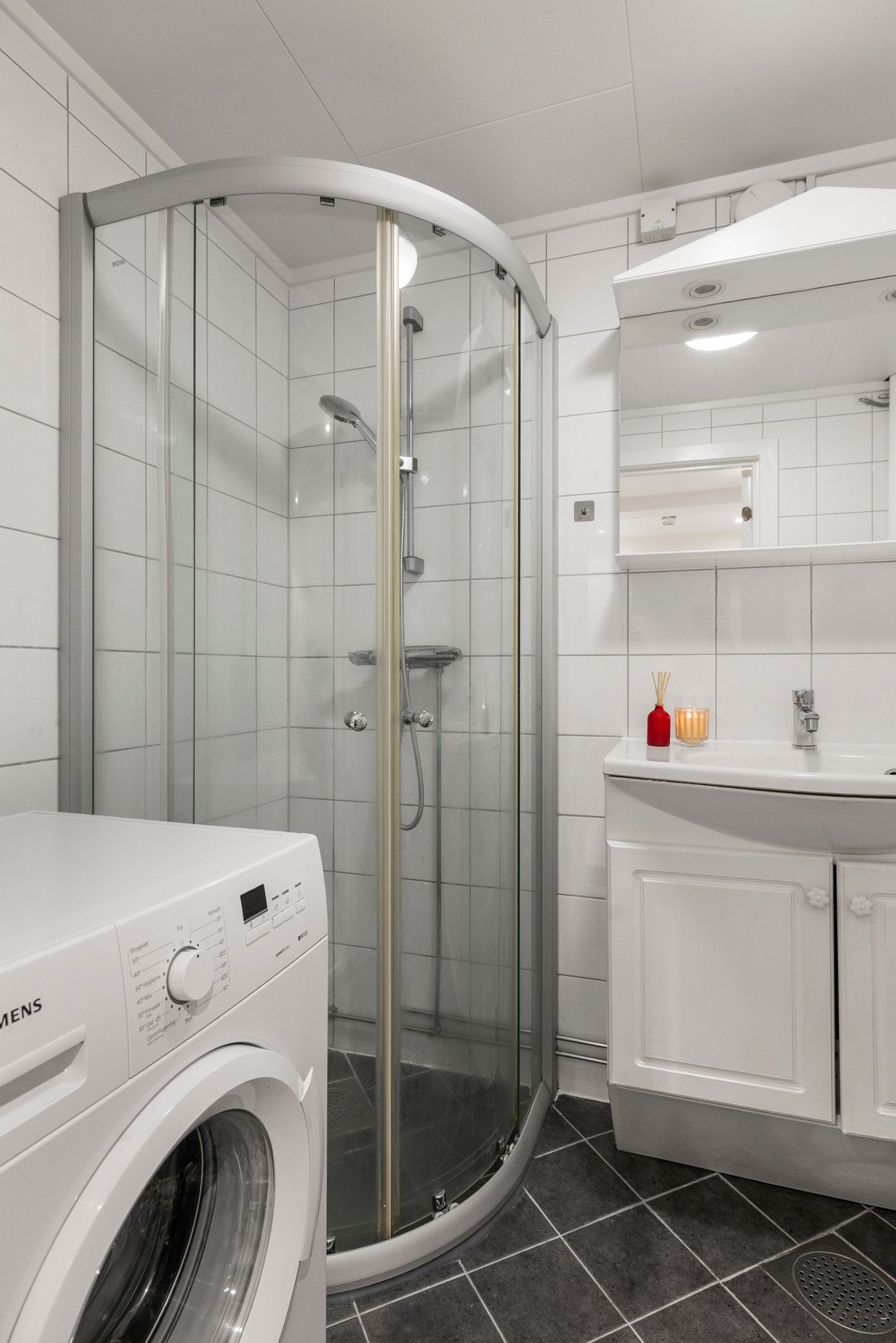 Rommet inkluderer en servant med profilerte fronter, gulvstående toalett og dusjhjørne, samt opplegg for vaskemaskin Galleribilde