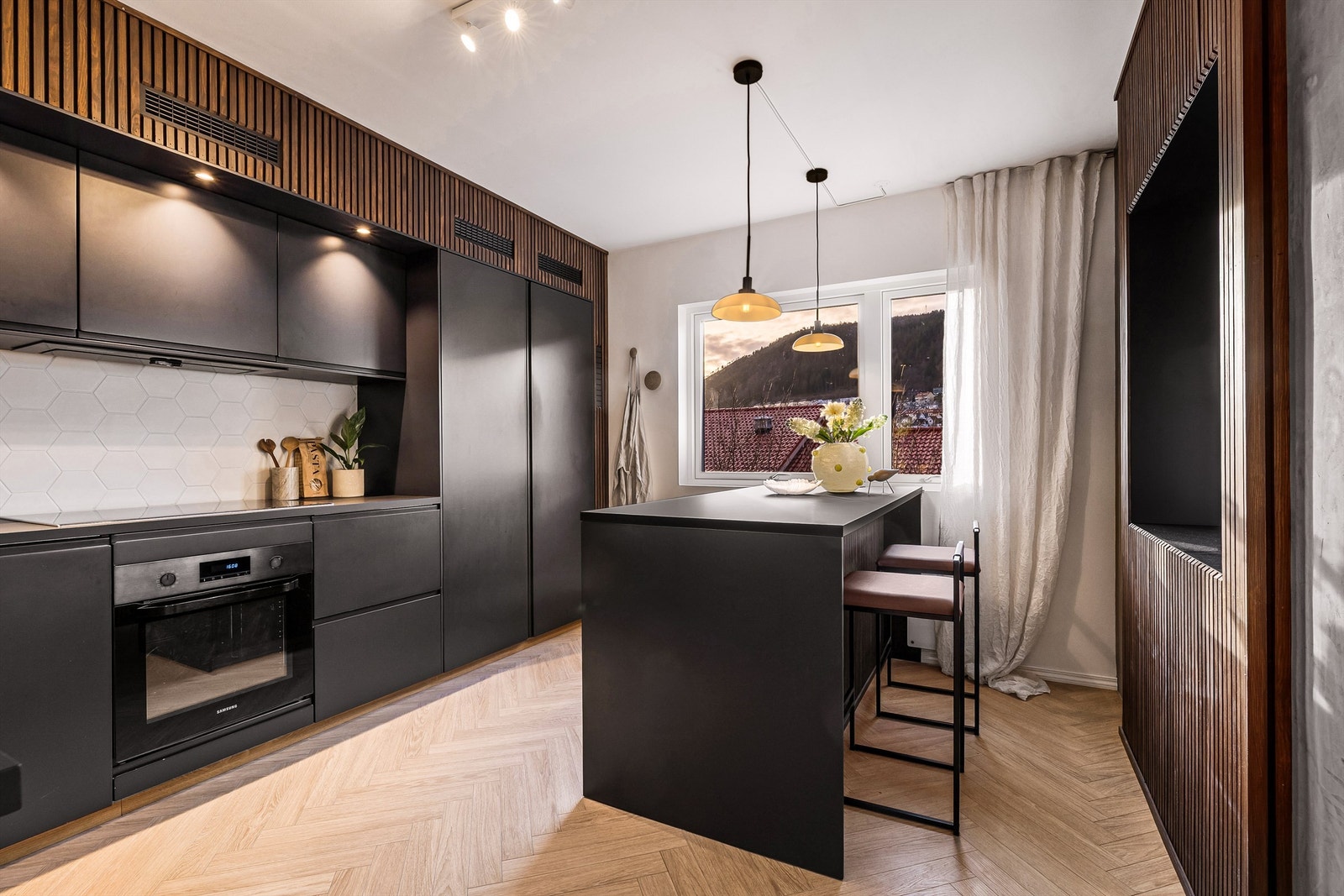 - Lekkert kjøkken med øy med spiseplass og store vindusflater. Legg merke til at spilene er i en mørkere tone på kjøkkenet, som passer perfekt til det slette, mørke kjøkkenet. - Galleribilde