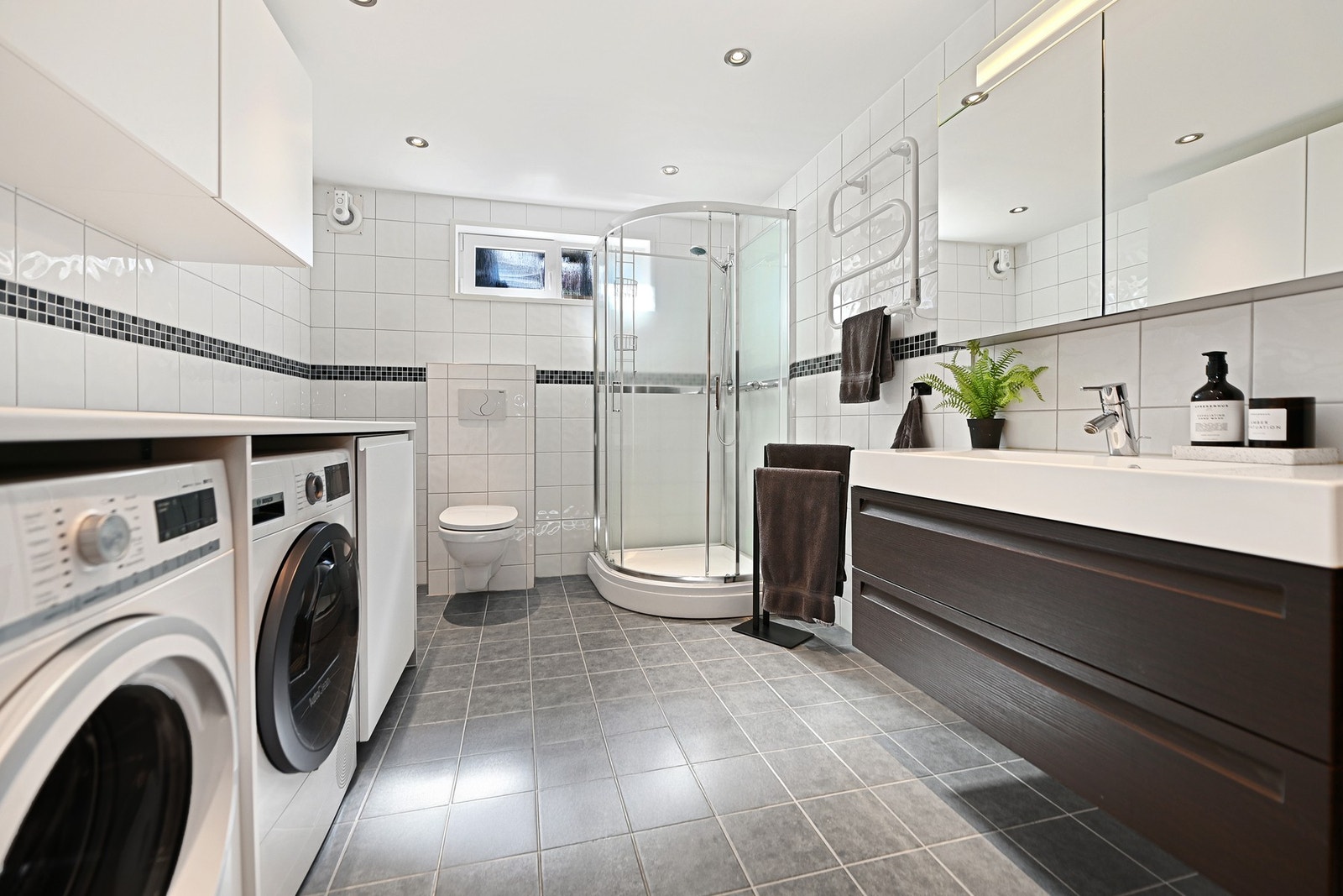 Badet i kjelleren er fra 2010 og fyller også behovet for vaskerom. Badet er innredet med nedfelt servant, veggfestet toalett, dusjkabinett og opplegg for vaskemaskin. Badet i kjelleren har ny vaskeromsinnredning. Galleribilde