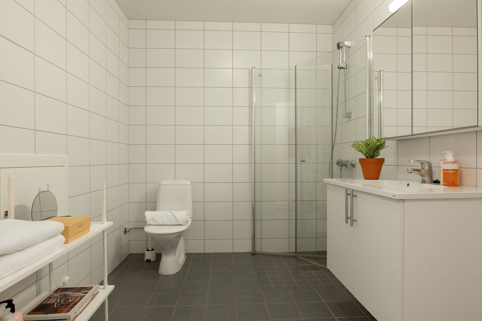 Lyst og delikat flislagt bad. Badet er holdt i lyse og tidløse farger med hvite fliser på veggene og mørke fliser på gulvet. Rommet er utstyrt med et praktisk dusjhjørne med innfellbare glassvegger, toalett og en romslig servantinnredning med speilskap. Galleribilde