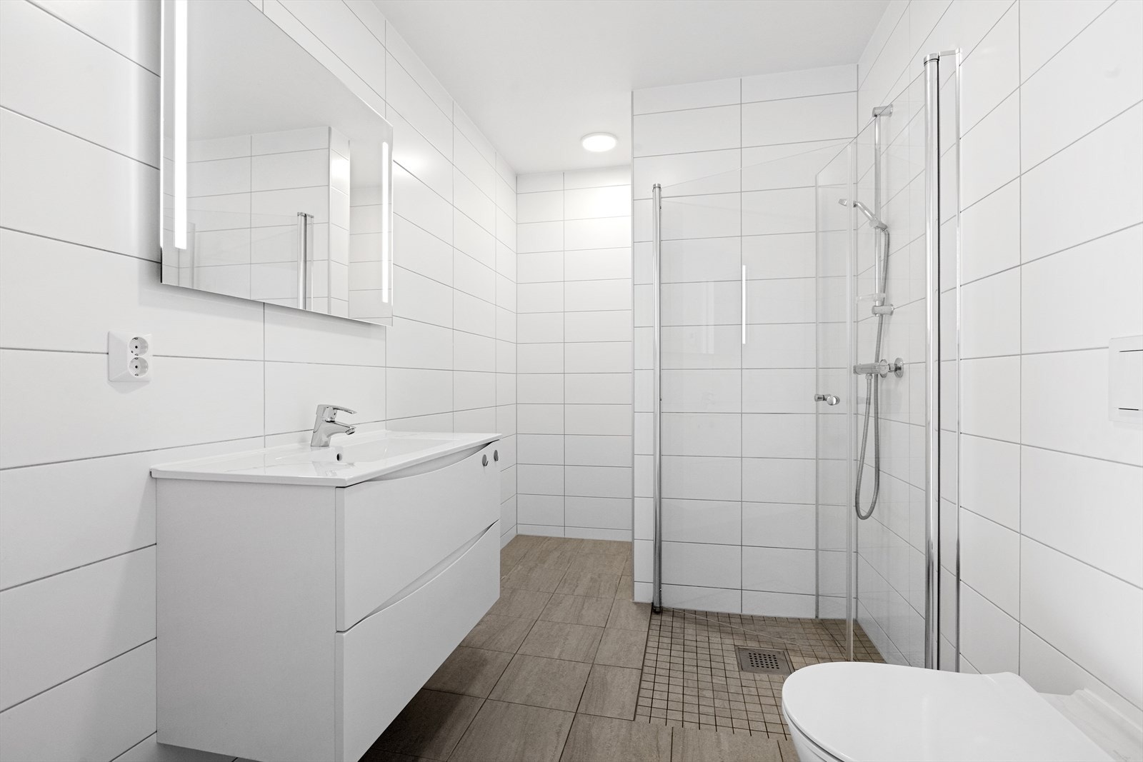 Flislagt bad/wc/vaskerom med varmekabler i gulv. Nedsenket dusjhjørne med glassvegger og veggmontert wc Galleribilde