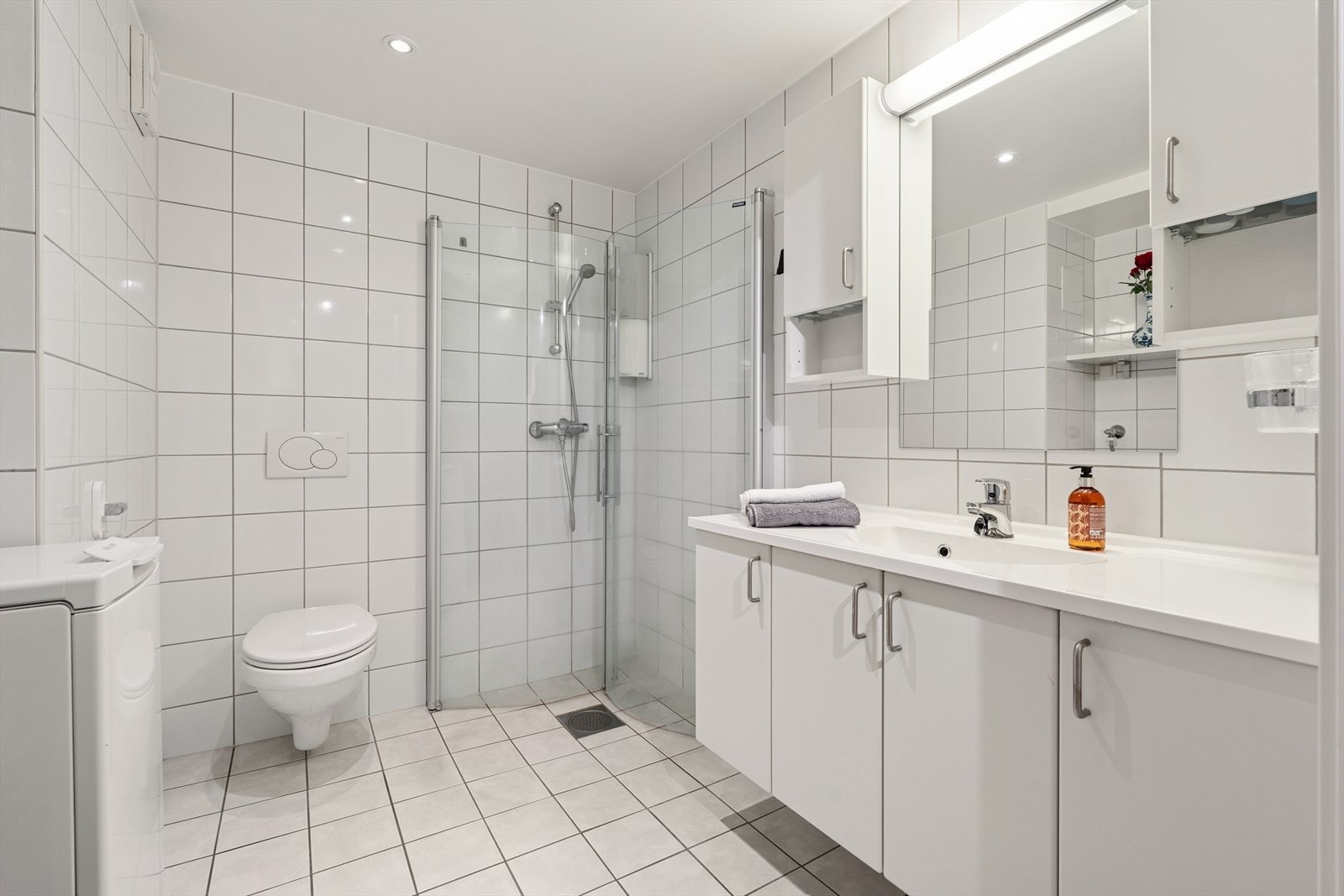 Lys og pent bad utstyrt med dusjhjørne, vegghengt toalett og opplegg for vaskemaskin. Galleribilde