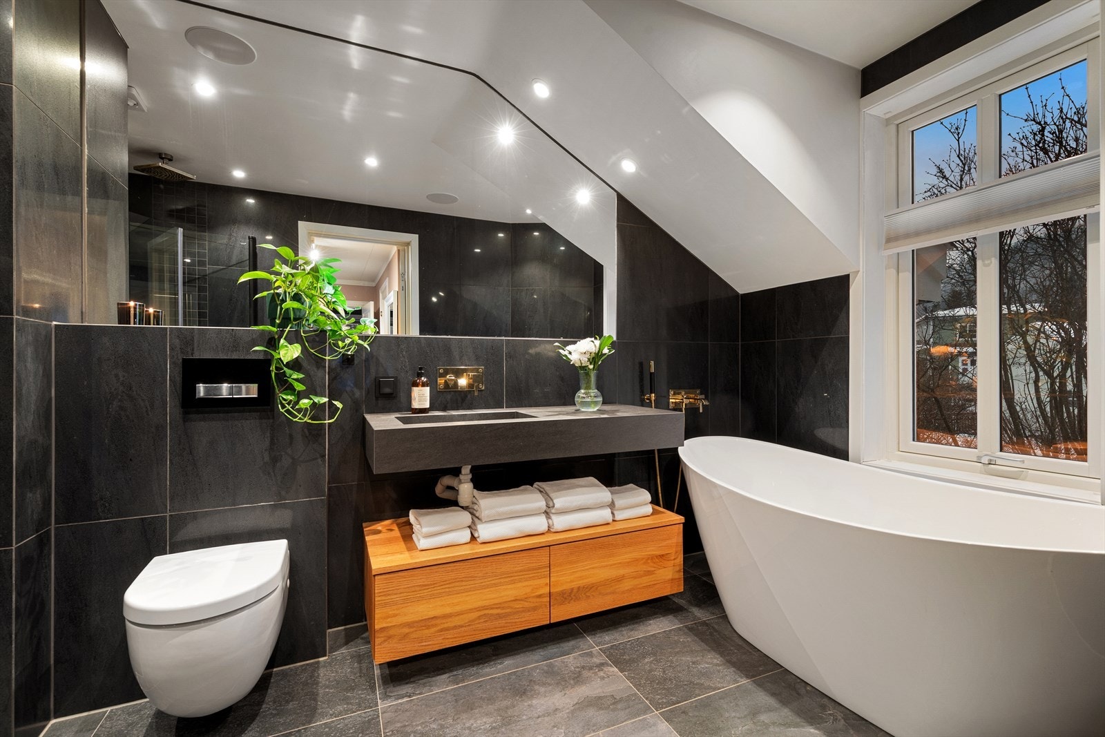 Badet inneholder dusjnisje med innfellbare dører, lekkert frittstående badekar, vegghengt toalett og delikat servant montert i benkeplate Galleribilde