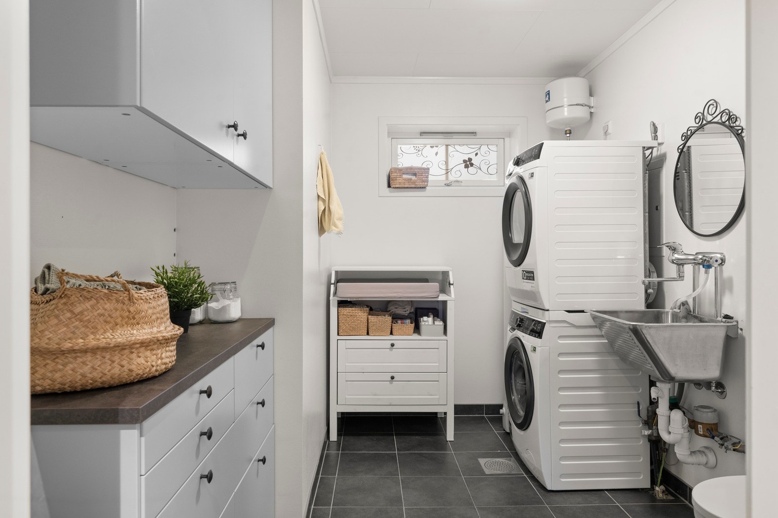 Praktisk vaskerom med flislagt gulv og sokkel, malt strie på vegger og hvite plater i tak som gir et lyst og ryddig preg. Rommet har varmekabler i gulvet og belysning i tak, samt utstyr med skyllekar, vegghengt toalett, varmtvannstank og opplegg for vaske Galleribilde