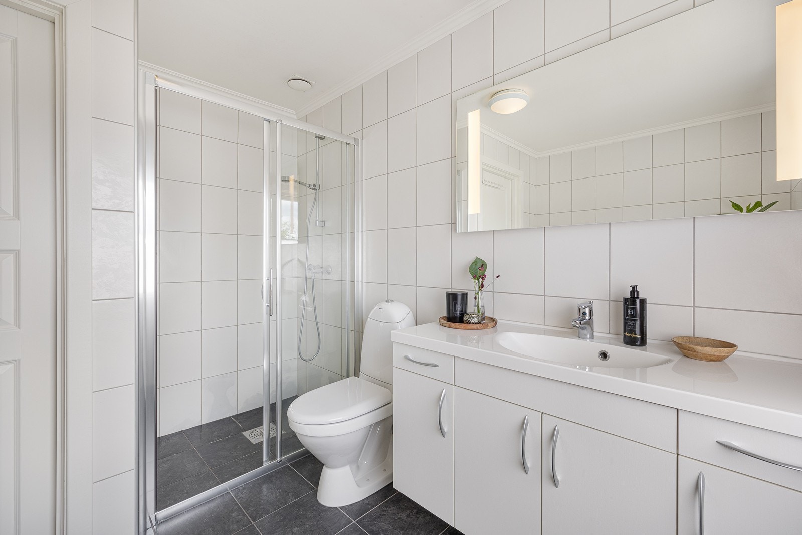 Innredningen har heldekkende servant over skapmøblement - i overkant er det speil med integrert belysning. Videre har badet dusjnisje med skyvedør og gulvmontert toalett. Galleribilde