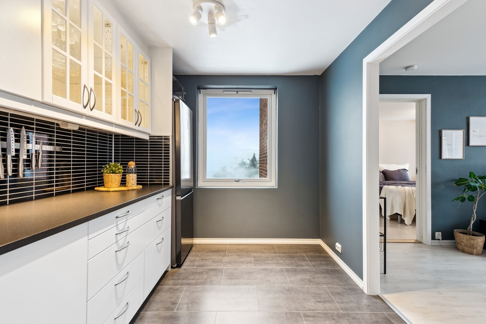 Delvis åpen løsning mellom stue og kjøkken. Det er lys i og under overskap, samt naturlig lys fra vindu. Galleribilde