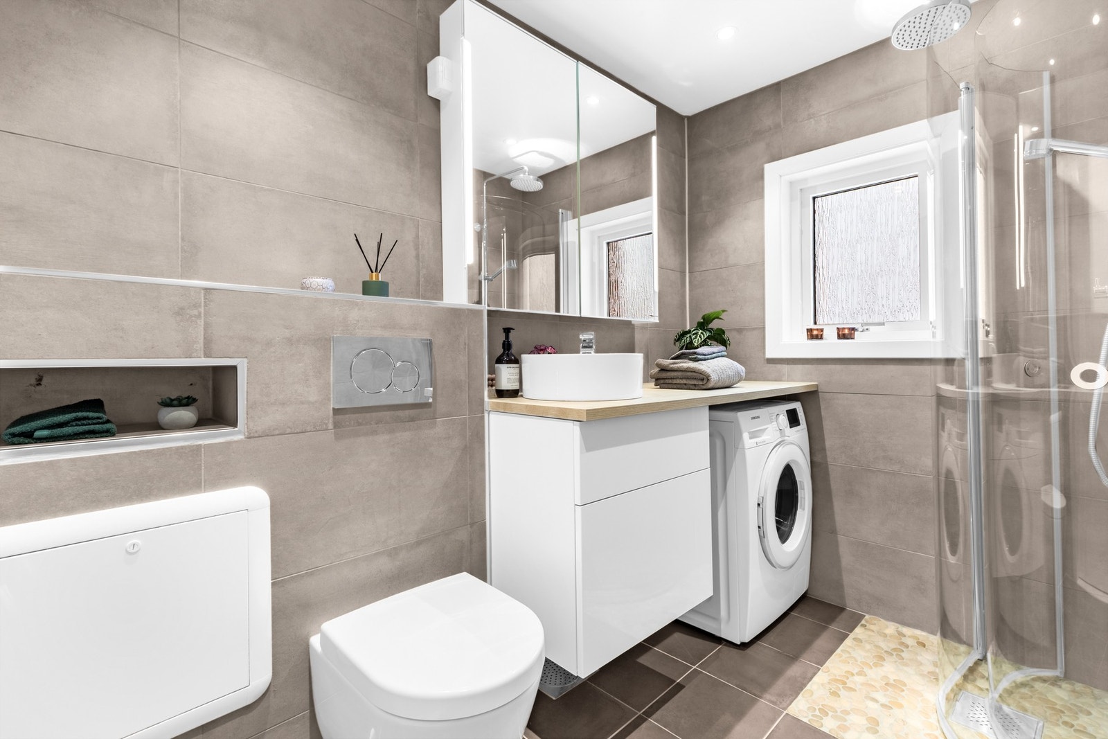 Et moderne bad som er utstyrt med varmekabler på gulv, downlights i tak, opplegg for vaskemaskin, vegghengt klosett, dusjesone med rullestein m.m. Galleribilde