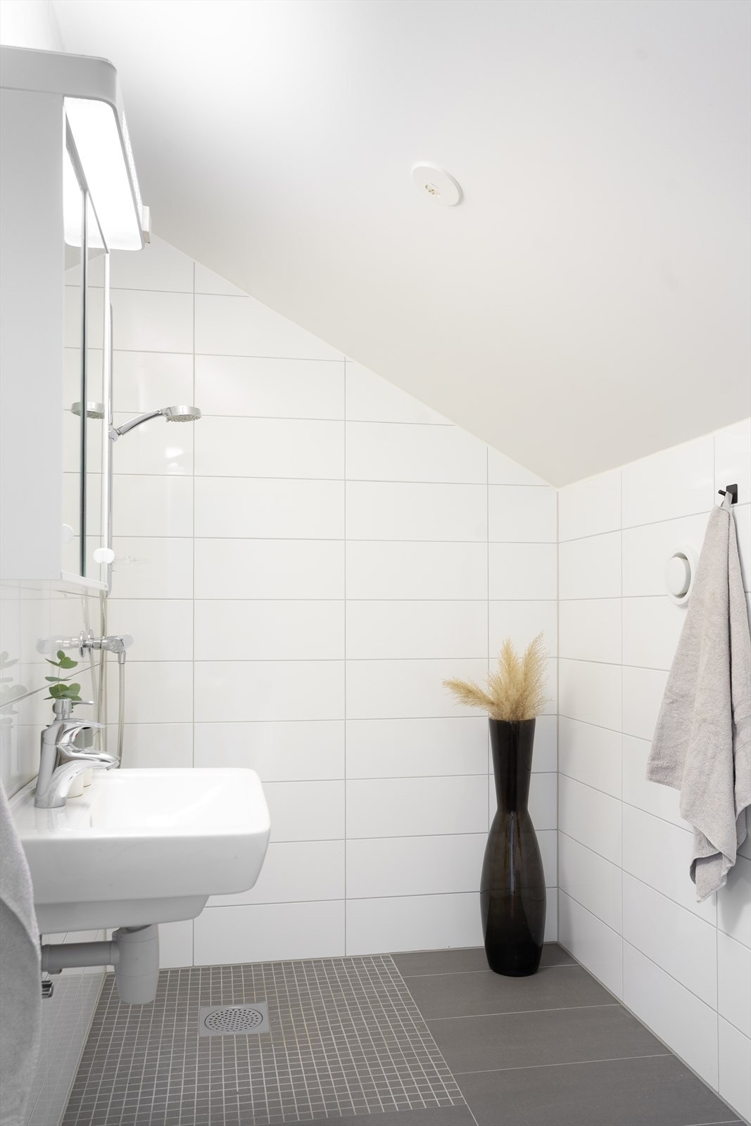 Pent bad på loftet med fliser på vegger og gulv samt mosaikkfliser i dusjsonen - badet er innredet med vegghengt toalett, speilskap, enkel servant og dusjsone med dusjgarnityr på vegg Galleribilde