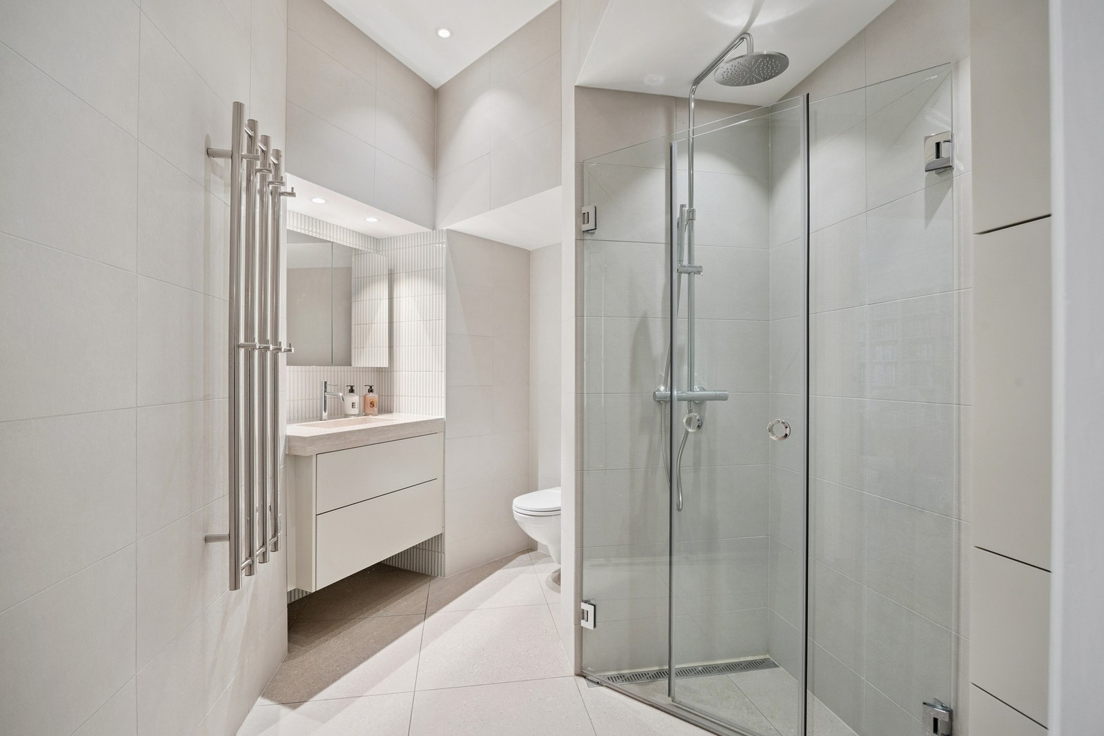 Badet med tilkomst fra entré er moderne innredet med gjennomgående flislagte gulv- og veggflater i lyse, tidløse toner. Galleribilde