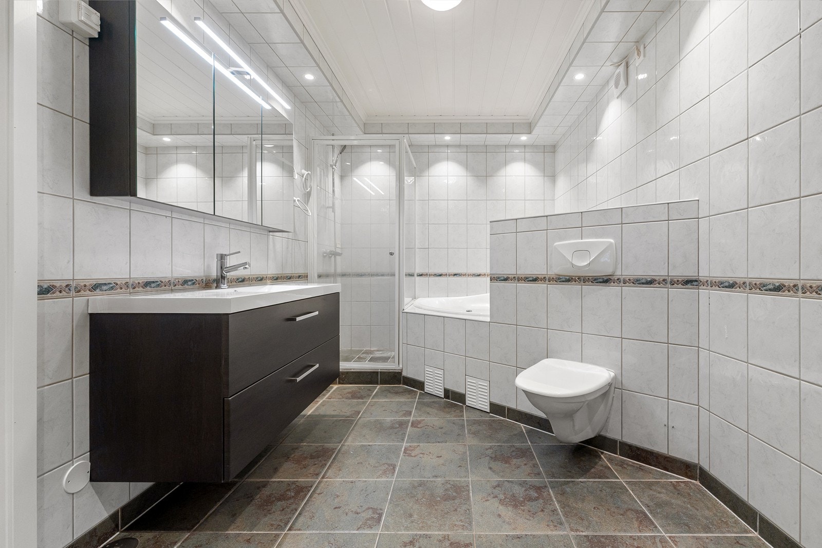 Bad med med gulvvarme, vegghengt servantskap, et vegghengt speilskap med belysning, dusjhjørne med opphøyet dusjsone og glassdør, innmurt boblebadekar og vegghengt toalett. Galleribilde