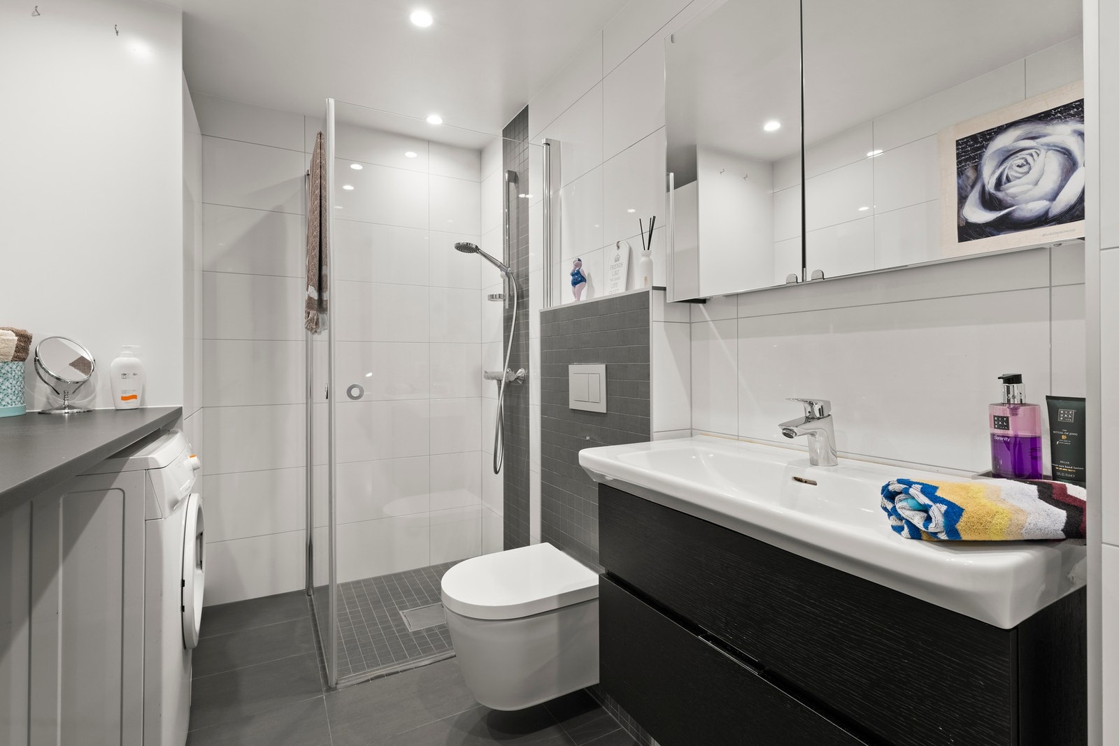 Det er flislagte overflater med varmekabler i gulv, og innredning med servant og underskap, speilskap med lys, vegghengt toalett og dusjhjørne med glassvegger. Galleribilde