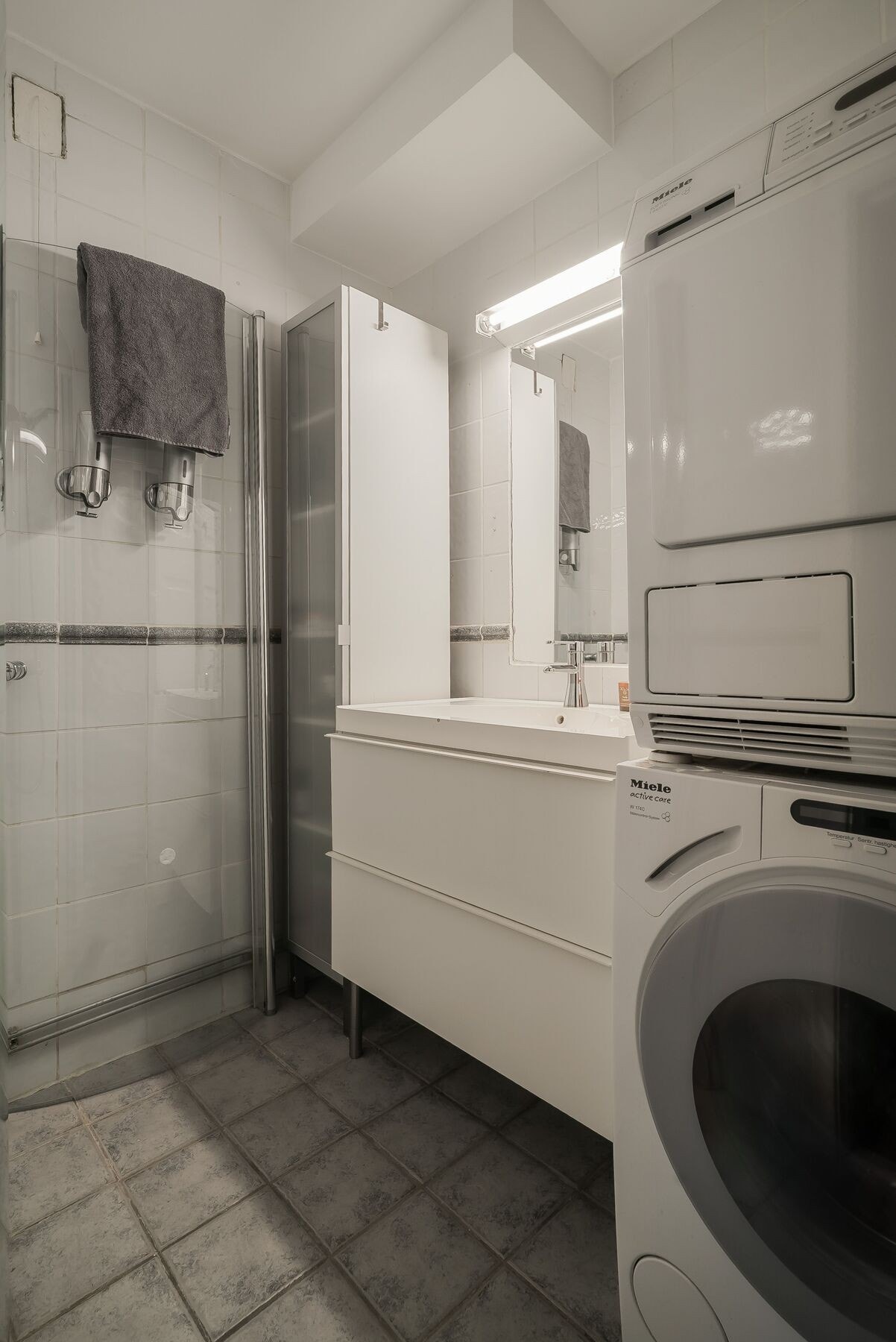 Bad på ca. 3,2 m² med flislagte overflater, dusjhjørne, servant med skuffer og opplegg for vaskemaskin. Galleribilde