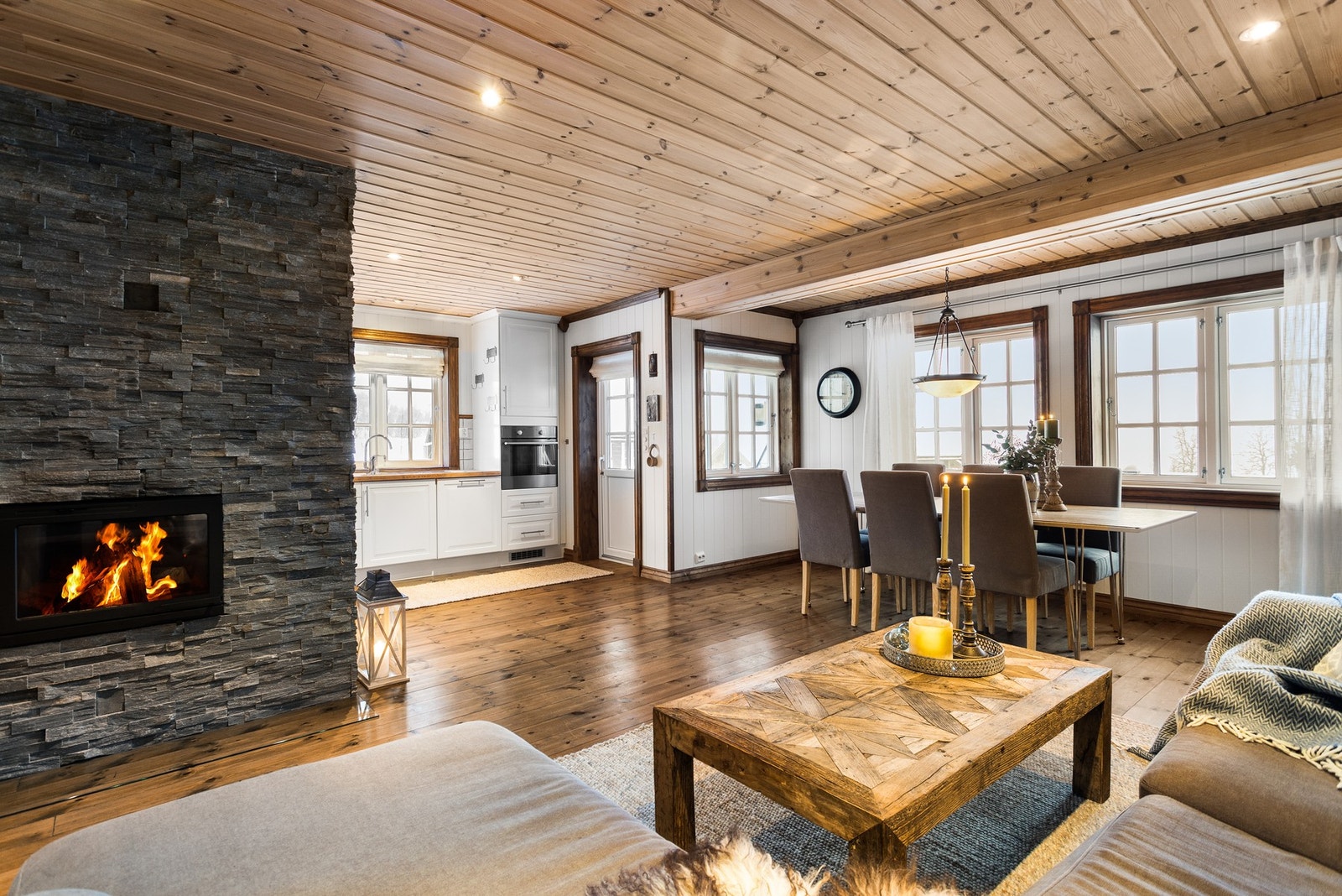 Romslig stue og kjøkken i åpen løsning. Midt i rommet står en flott peisovn med pipe forblendet i skiferpanel Galleribilde