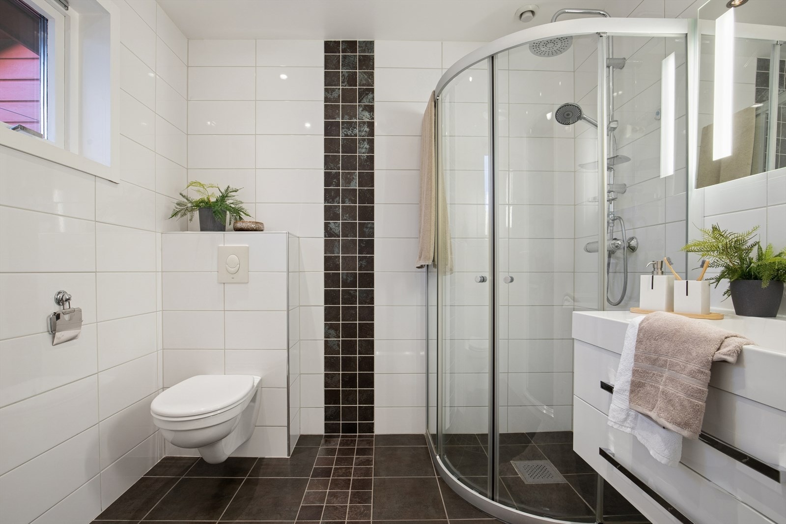 Flislagt baderom bestående av speil med belysning, servant med seksjon, veggmontert toalett og dusjhjørne. Galleribilde