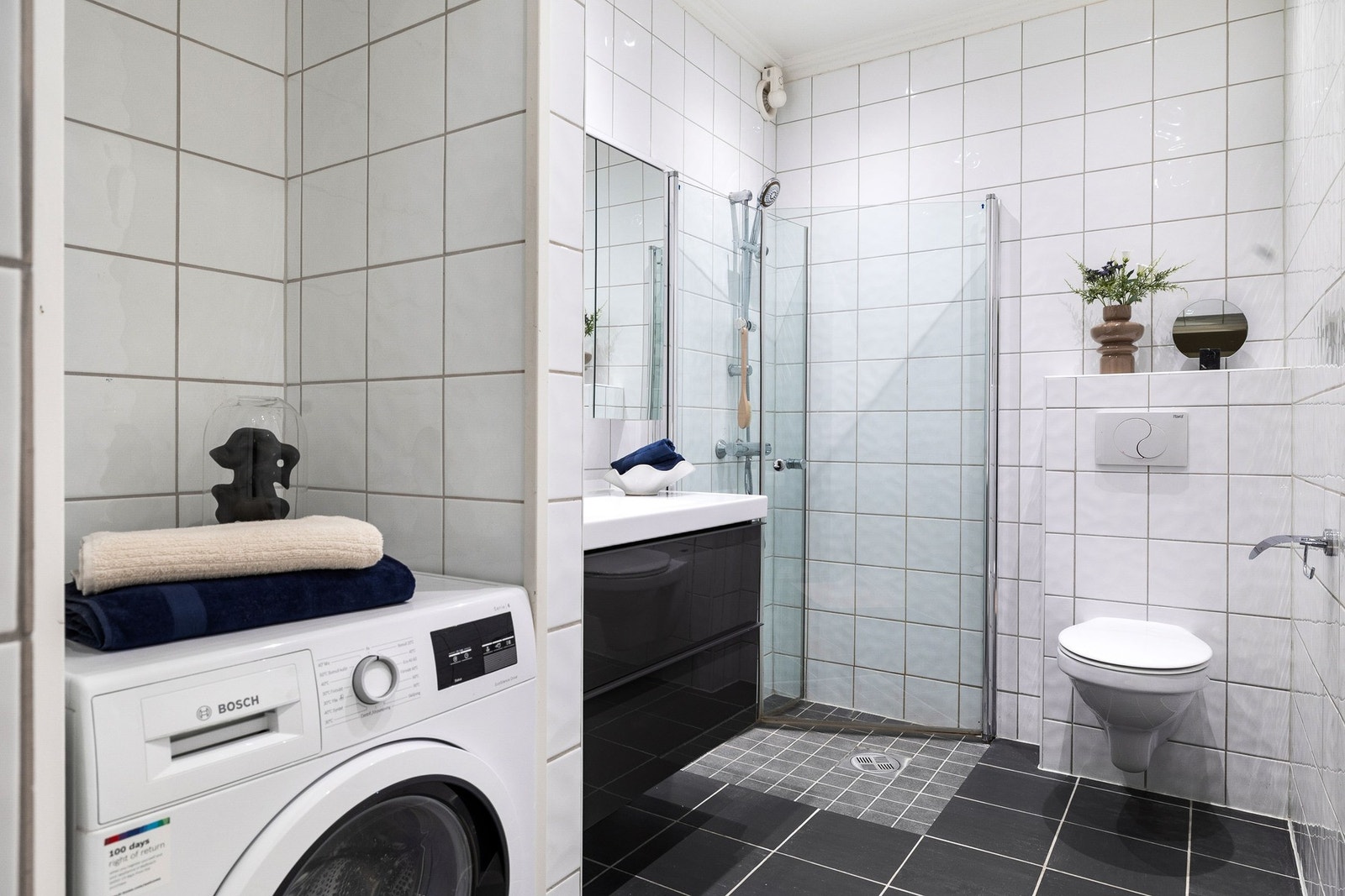 Flislagt baderom med praktisk innredning med vask, dusj, vegghengt toalett og opplegg for vaskemaskin. Galleribilde