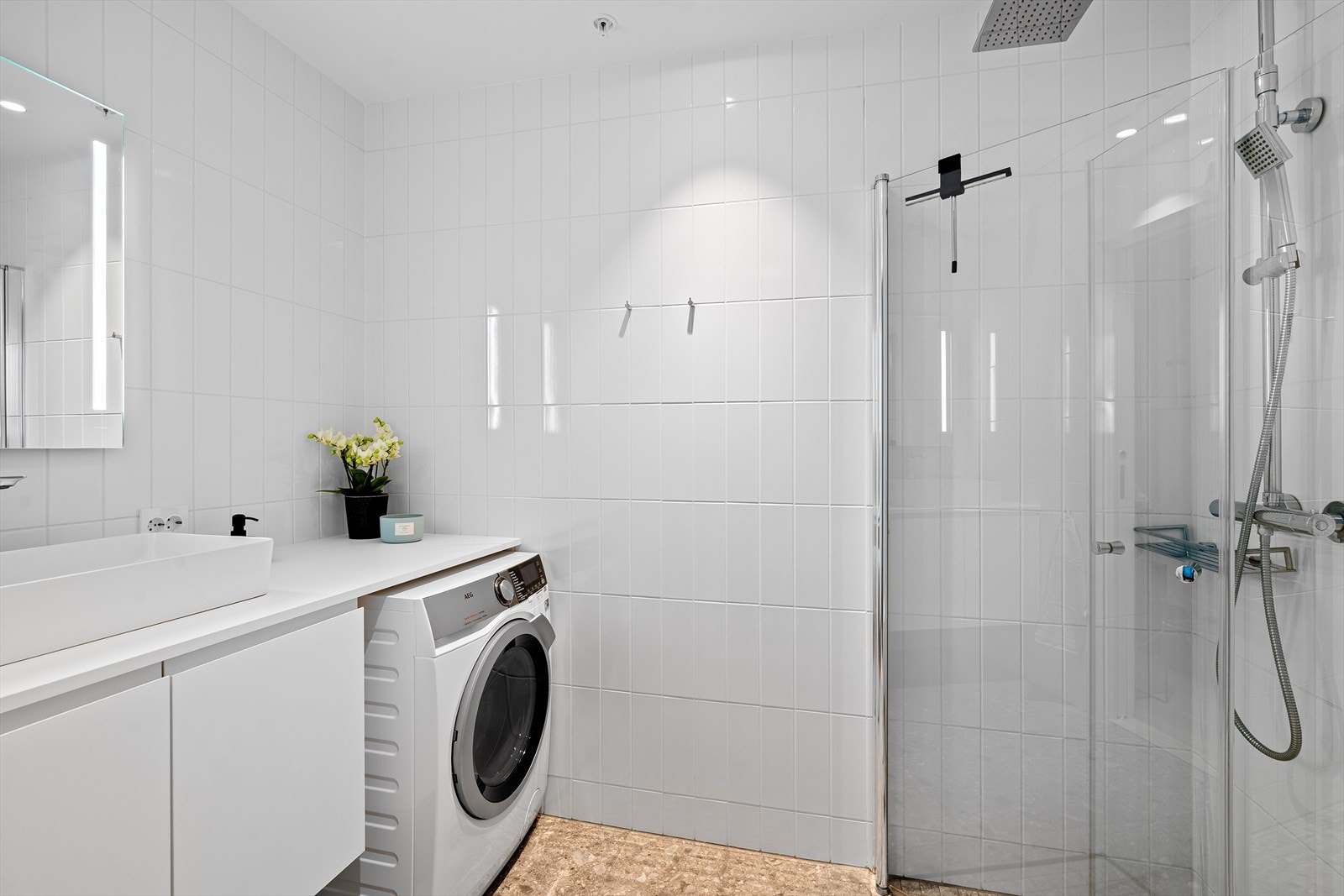 Badet er helfliset med varme i gulvet. Det inneholder en baderomsinnredning med benk over vaskemaskin, speil med innfelt belysning, vegghengt toalett, samt dusjnisje med regndusj og innfellbare dusjdører. Galleribilde