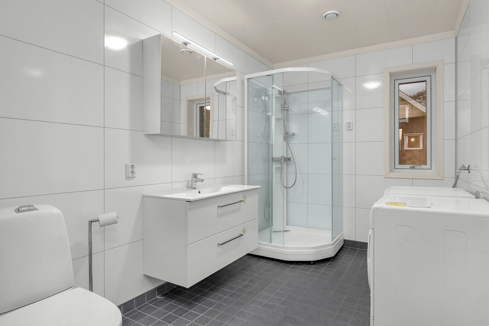 Bad/vaskerom har flislagt gulv med gulvvarme og våtromsplater på veggene. Rommet er innredet med servant på skuffeseksjon, speilskap med lys, dusjkabinett, wc og opplegg for vaskemaskin. Galleribilde