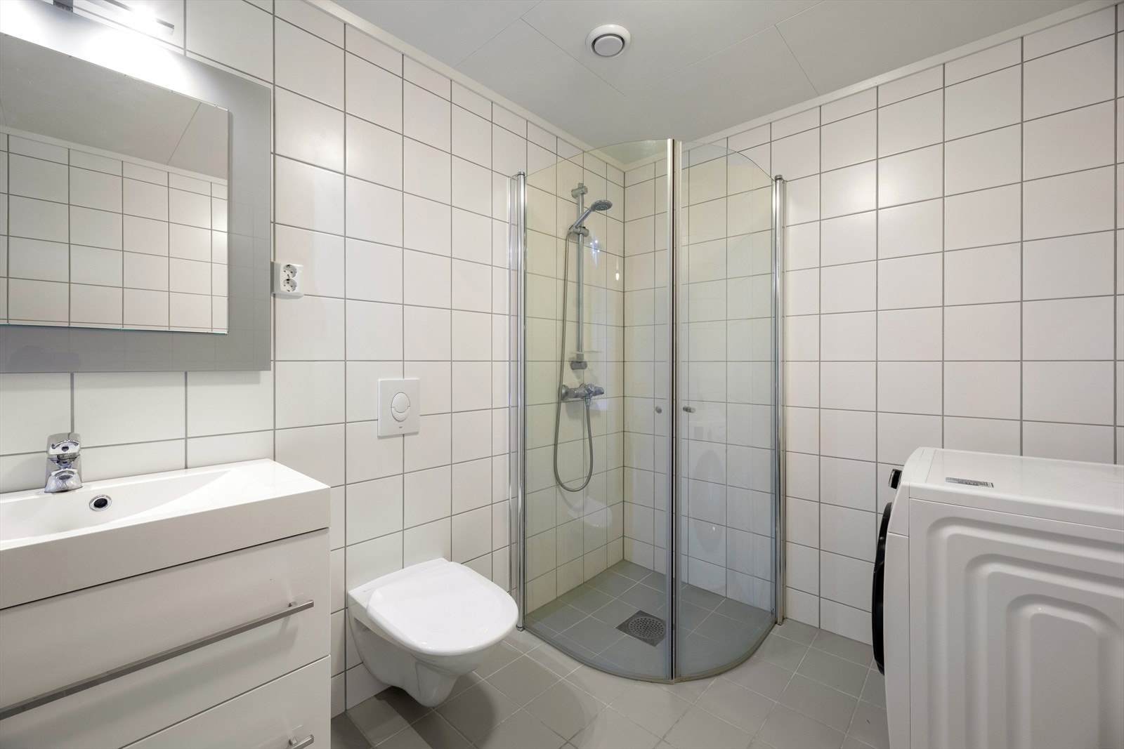 Badet er ellers utstyrt med dusjhjørne, vegghengt toalett og baderomsinnredning med nedfelt servant. I tillegg har man opplegg for vaskemaskin og plass til tørketrommel. Avtrekk via mekanisk vifte. Galleribilde