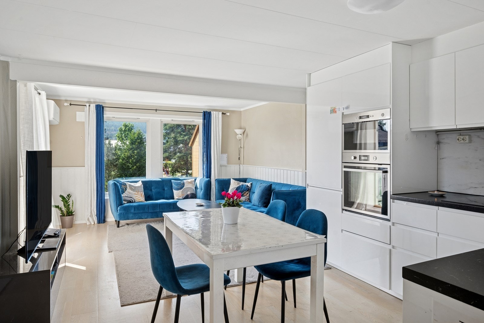 Pent kjøkken med slette hvite fronter med integrerte hvitevarer som stekeovn, mikrobølgeovn, kjøleskap, oppvaskmaskin og platetopp. Galleribilde