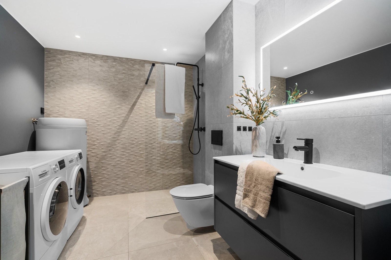 Varmekabler i gulv, vegghengt toalett, stor dusj. Opplegg for vaskemaskin samt plass til tørketrommel. Grå fliser på gulv og vegg, glassvegg i dusj. Galleribilde