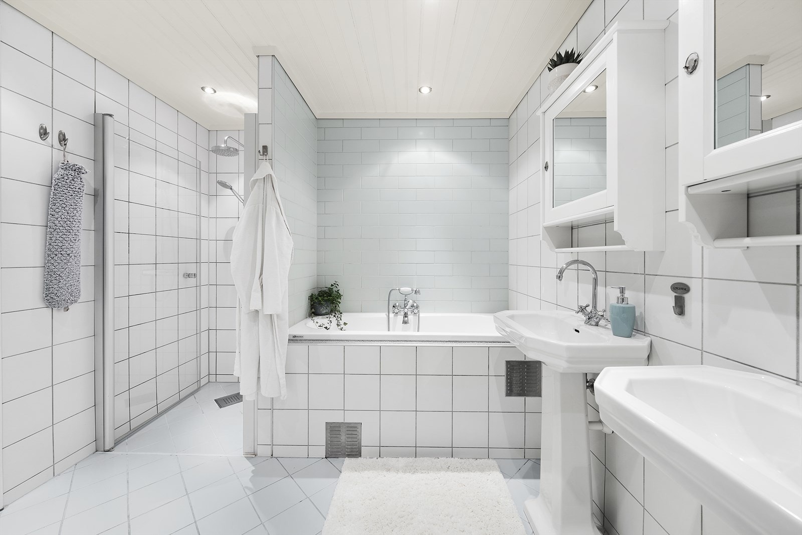 Badet er utstyrt med to servanter med overliggende speilskap, vegghengt skap, badekar, dusjhjørne med innfellbar glassvegg og vegghengt toalett Galleribilde