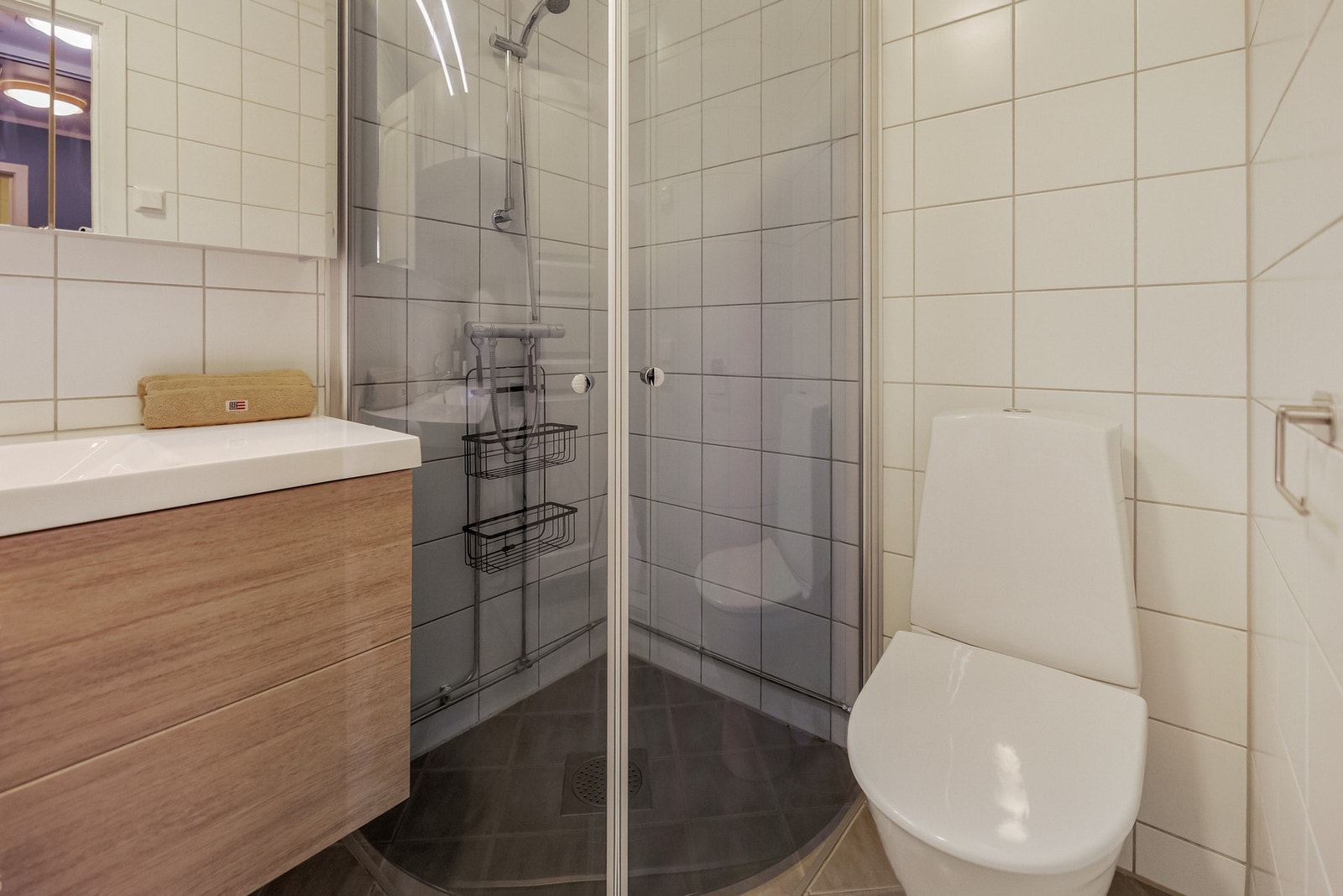 Rommet inneholder en vegghengt baderomsinnredning med servant, et gulvstående toalett og et praktisk dusjhjørne med dører. Galleribilde