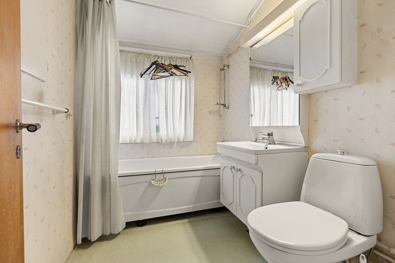 Lyst bad som er utstyrt med belegg på gulv og tapet på veggene. Det er også badekar med dusjforheng, gulvmontert WC, heldekkende servant og speil. Galleribilde