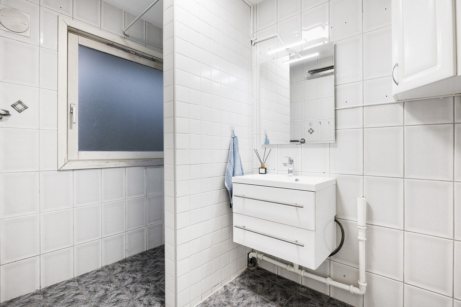 Flislagt bad med en innredning bestående av servant med skuffeseksjon, speil med overlys, åpen dusjløsning og opplegg for vaskemaskin. Galleribilde