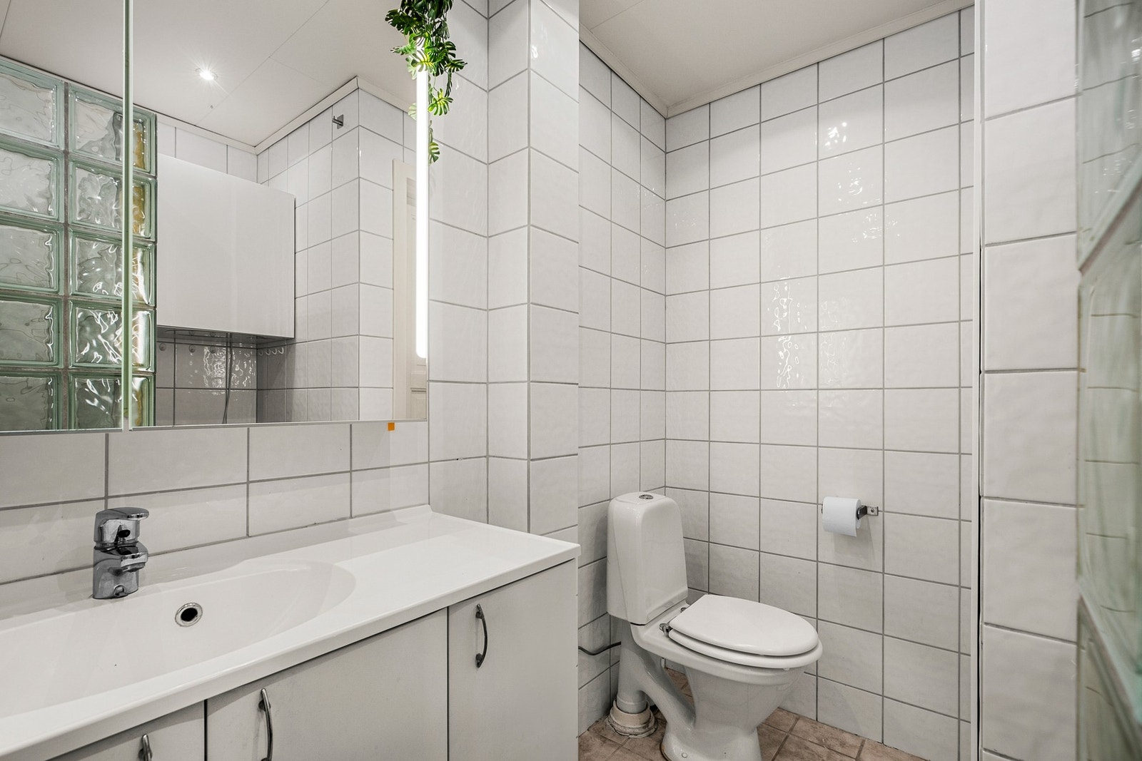 Badet er utstyrt med gulvmontert toalett, servantinnredning med veggmontert speil og integrert belysning, dusjhjørne med vegger i glassbyggerstein, opplegg for vaskemaskin Galleribilde