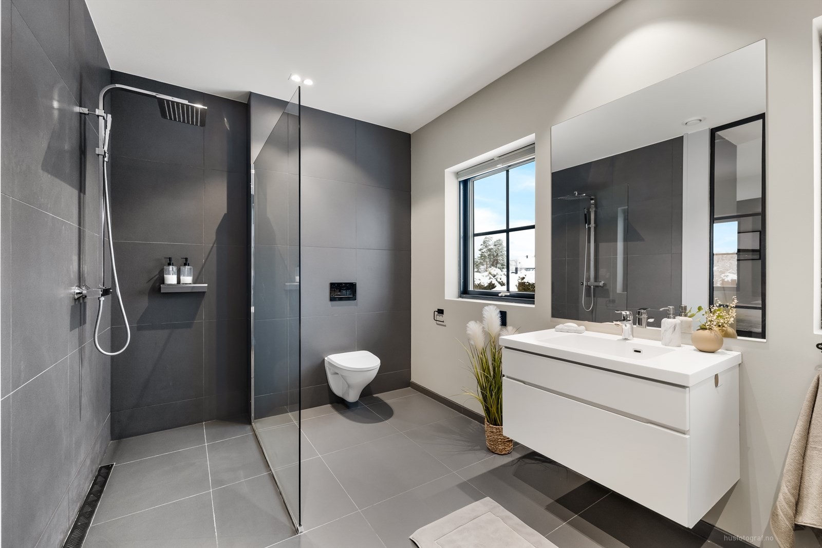 Delikat og innbydende flislagt bad med hvit baderomsinnredning med nedfelt servant, speil, vegghengt toalett og dusjsone med regndusjarmatur. Galleribilde