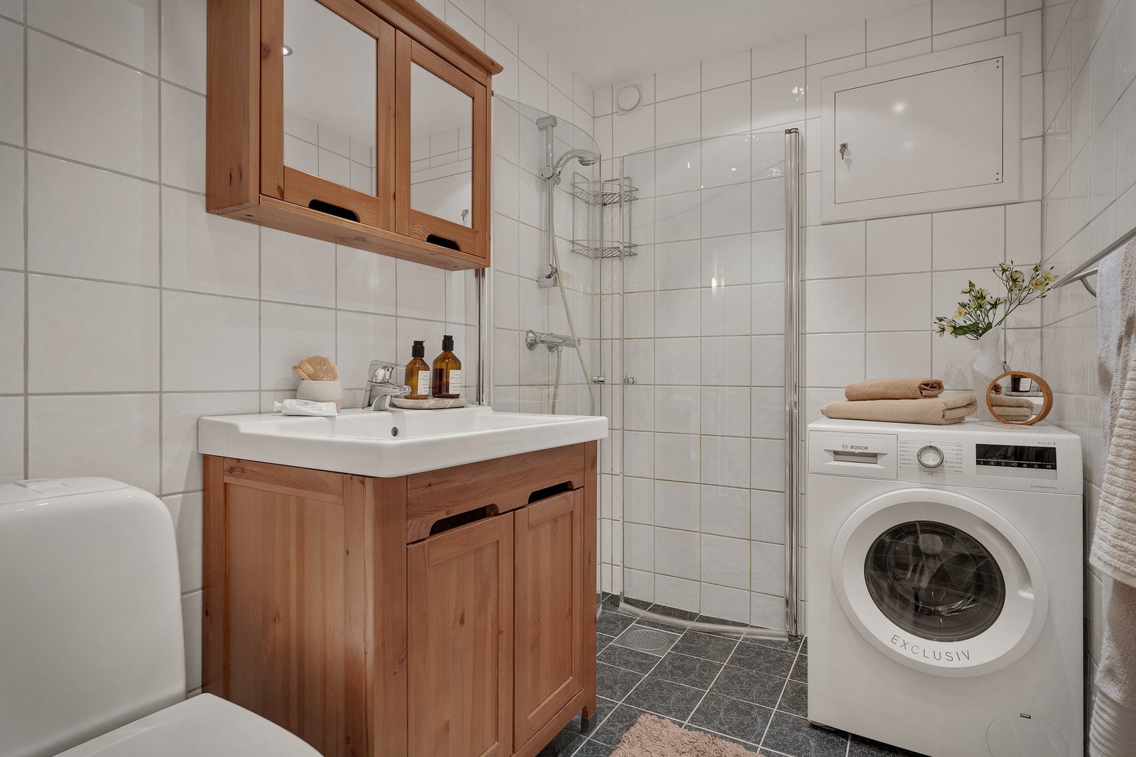 - Badet er lyst og klassisk, med fliser på gulv og vegger, toalett, servant med benkeskap, opplegg for vaskemaskin og dusjdører i glass. - Galleribilde