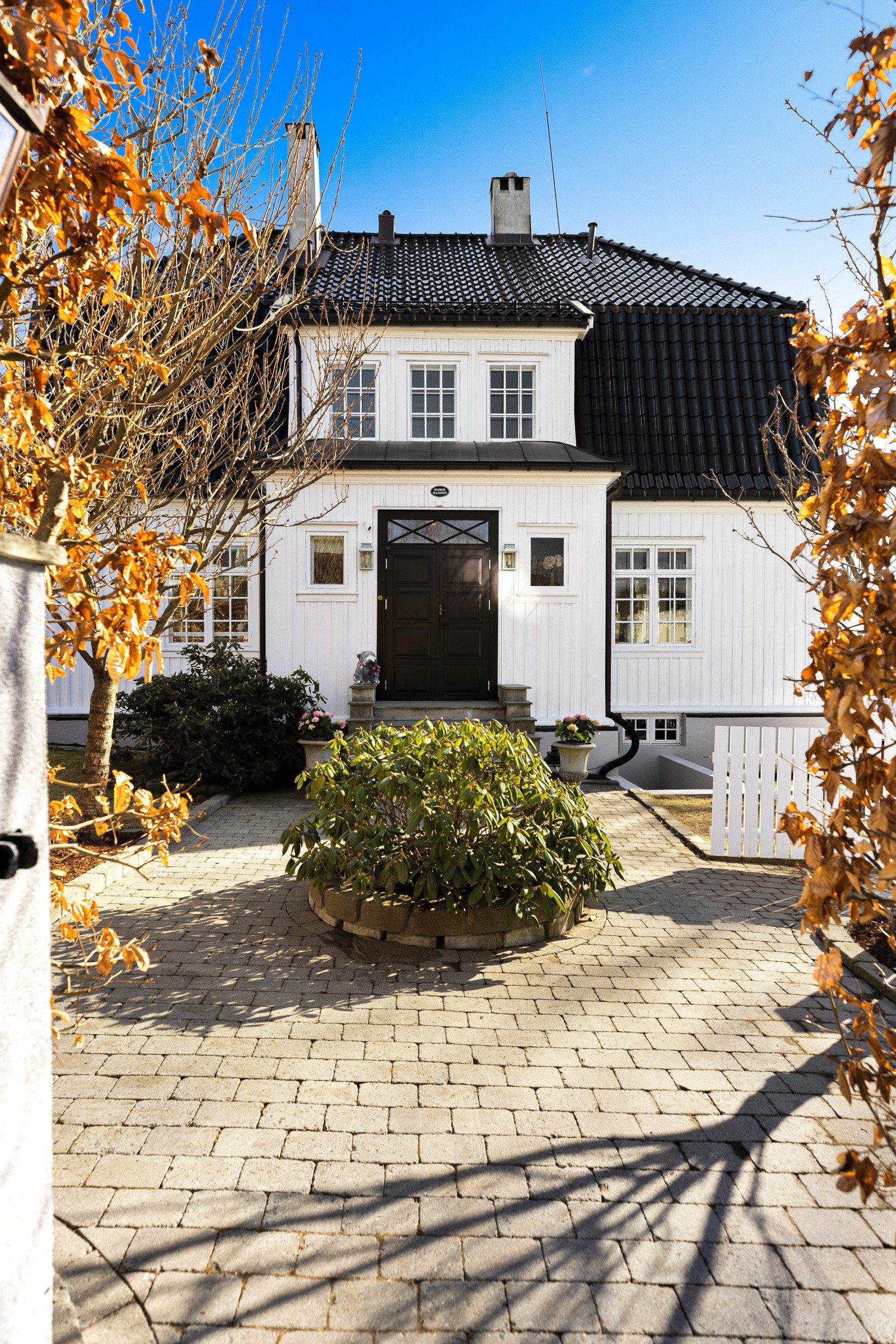 Boligen har adkomst via steinsatt gangareal med trær på sidene, smijernsport mot vei og hvit port mot felles gårdsplass.