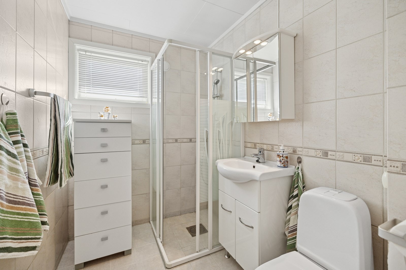 Baderommet er utstyrt med gulvstående klosett, dusjhjørne med veggmontert dusj og servant med skapdører under. Galleribilde