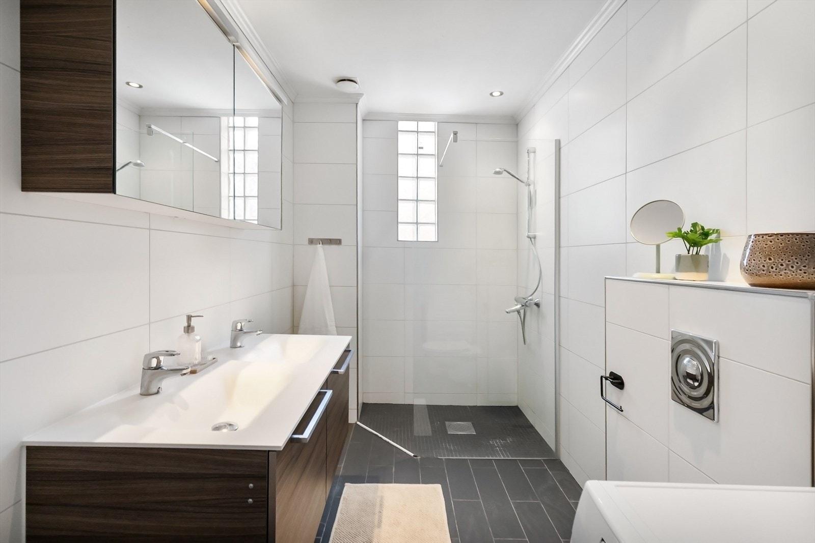 Badet fremstår som funksjonelt og delikat, med moderne fasiliteter og tidløse materialvalg som sikrer både komfort og stil. Dusjhjørne med herdet glass-skyvedører, dobbelservant og vegghengt toalett. Galleribilde