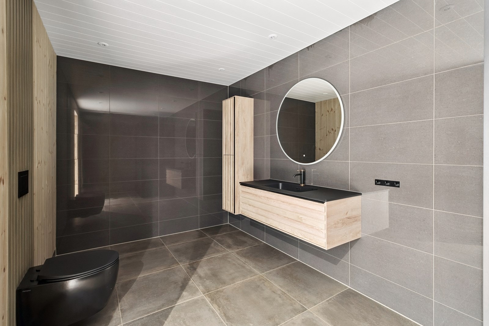 Et stort og moderne bad med store fliser og vegghengt toalett. Noe arbeid gjenstår. Galleribilde