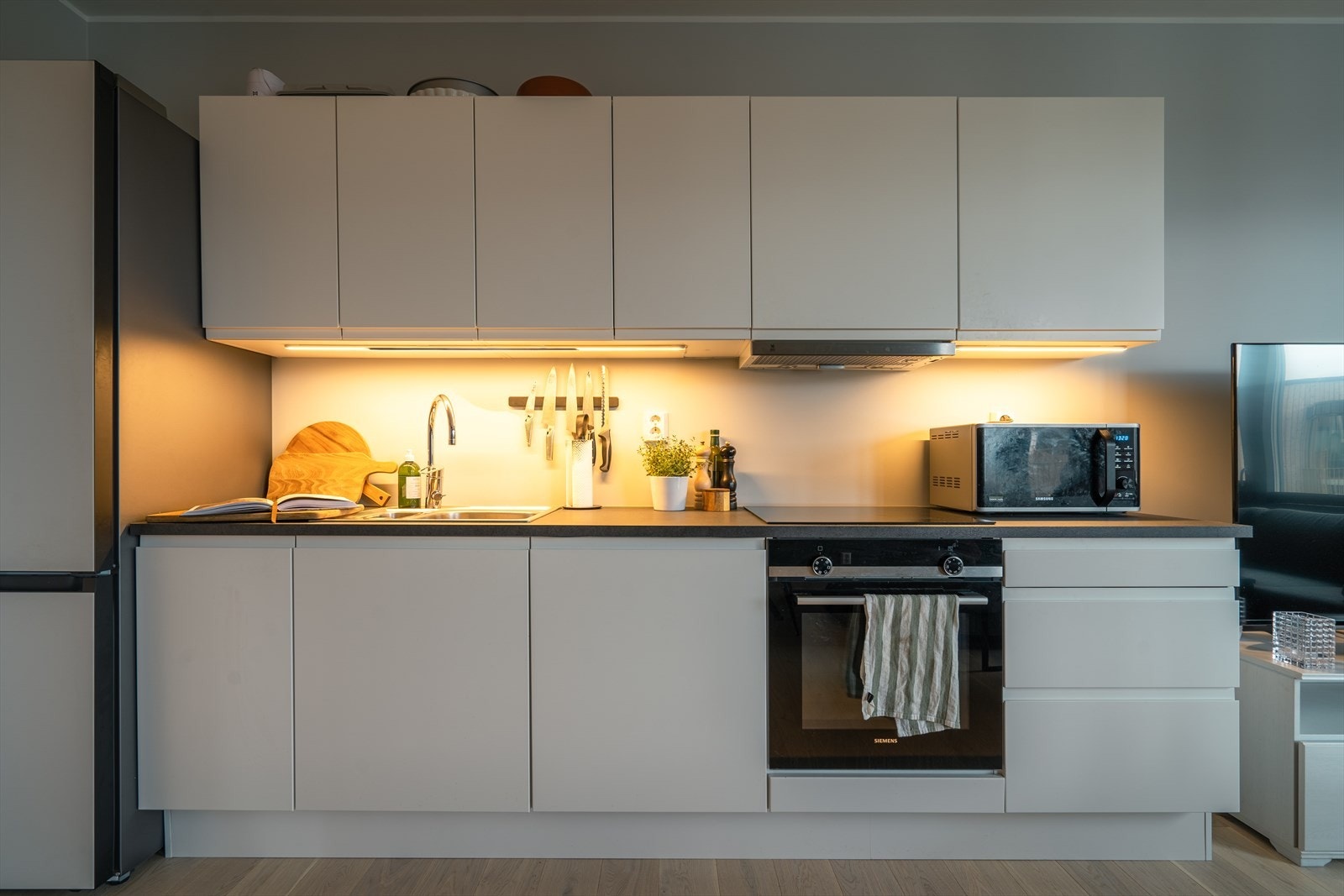 Det delikate kjøkkenet har alt man trenger plassert på én vegg. Innredningen har hvite, slette fronter og hvit laminat benkeplate med nedfelt dobbeltkum. Foto: Privat. Galleribilde