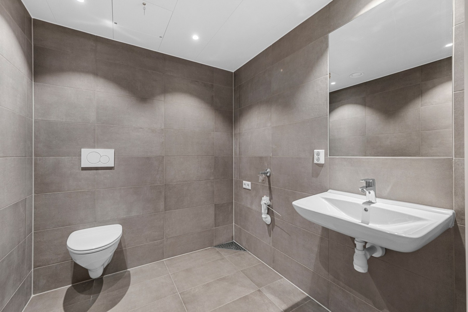 Stilig og flislagt vaskerom med gulvvarme, vegghengt wc, servant med speil og opplegg for vaskemaskin. Galleribilde