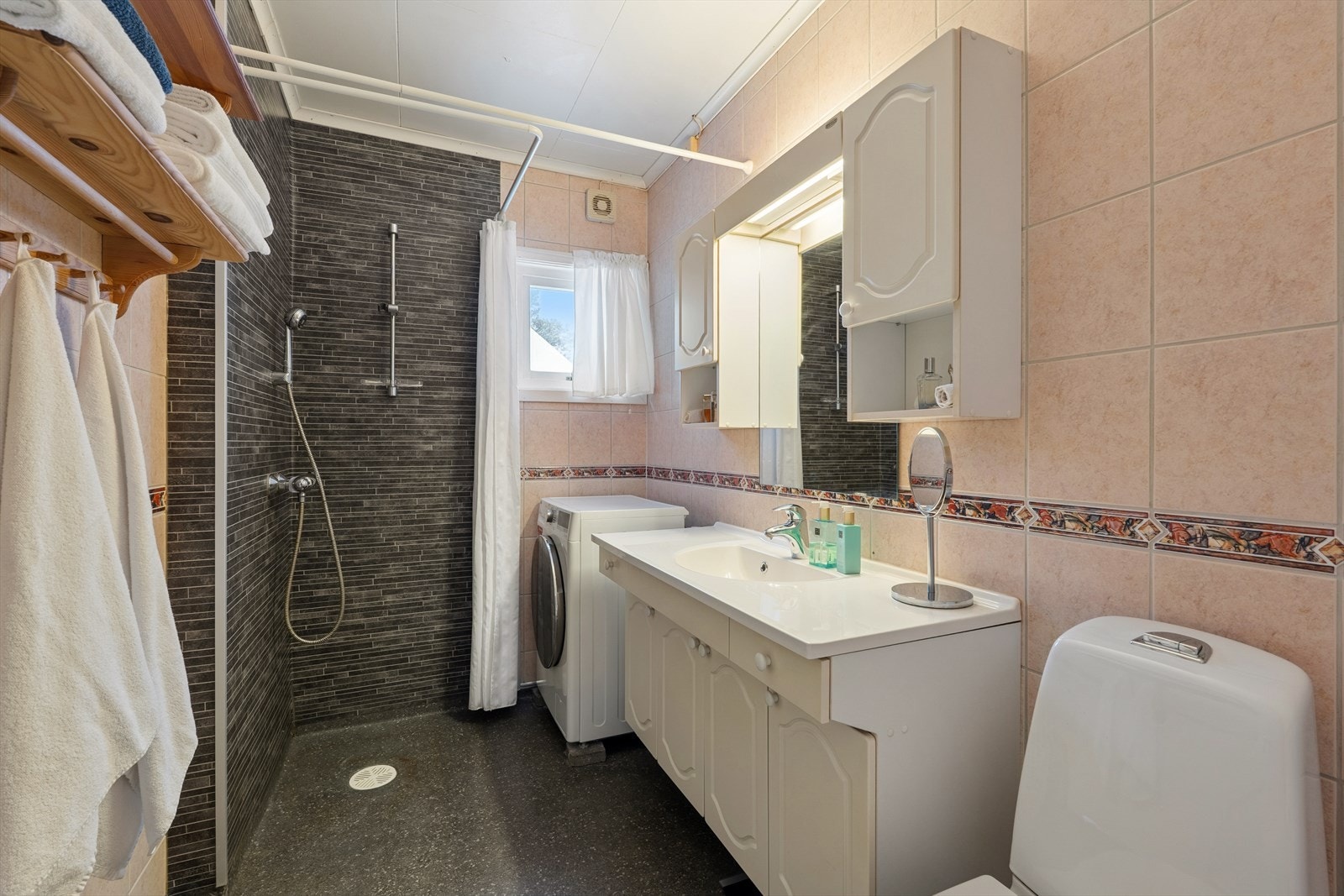 Baderom med dusjanretning på vegg, servant med skapinnredning, opplegg til vaskemaskin samt toalett. Galleribilde