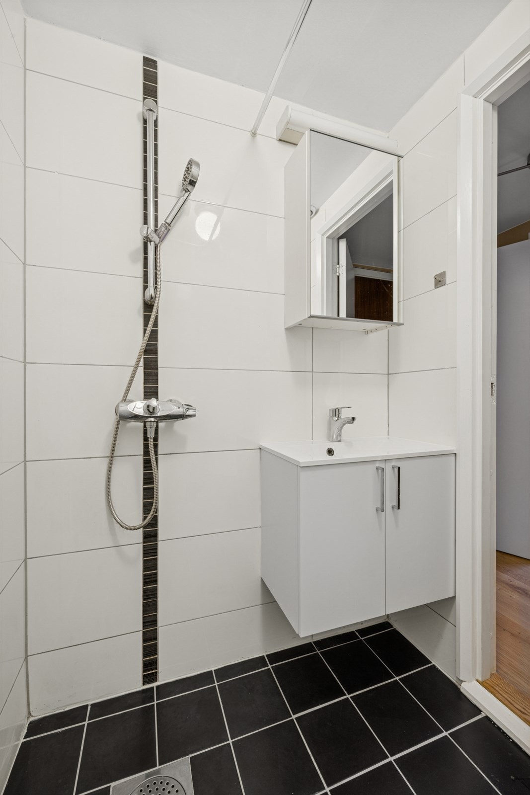 Rommet er innredet med en åpen dusj, servantinnredning med nedfelt servant i skap, og et veggskap med speilfront. Galleribilde