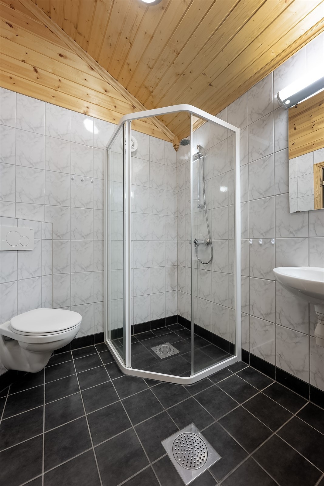 Badet har flislagt gulv med varmekabler, wc, dusjhjørne og servant med speil og lys Galleribilde