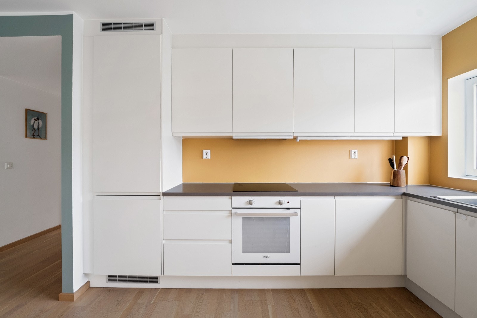 Kjøkkeninnredning med hvite, glatte fronter og mørk laminat benkeplate. På kjøkkenet er det integrert stekeovn, induksjonstopp, kjøl-/fryseskap, oppvaskmaskin, oppvaskkum. Ventilator over induksjonssoppen Galleribilde