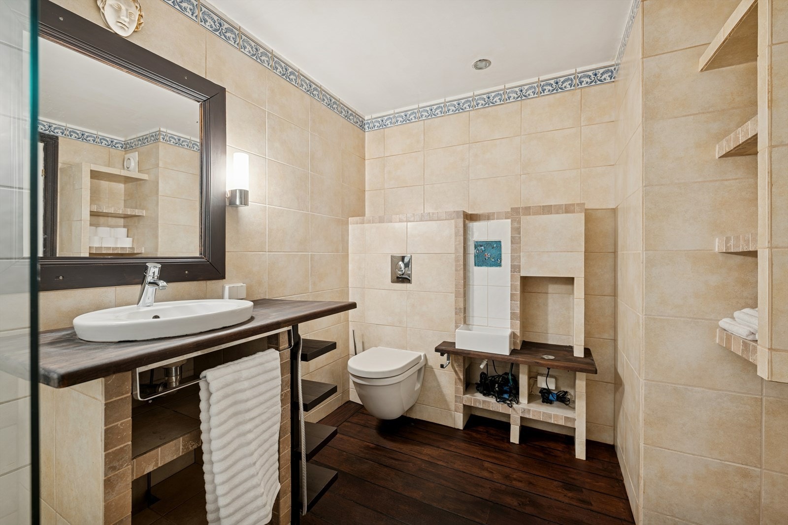 Eldre flislagt baderom som består av vegghengt klosett, nedsenket vask med hyller og dusjkabinett Galleribilde