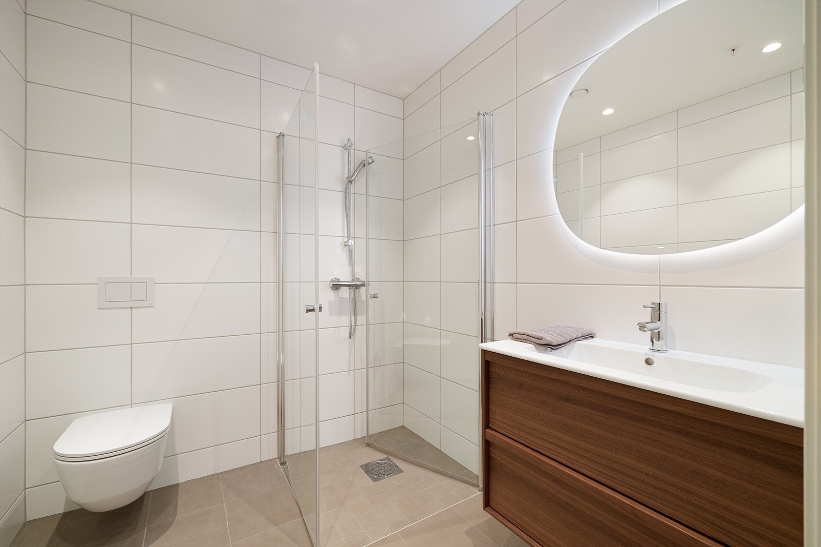 Badet inneholder servantmøbel, speil m/integrert belysning, vegghengt toalett og dusj med innfellbare dører i herdet glass. Galleribilde