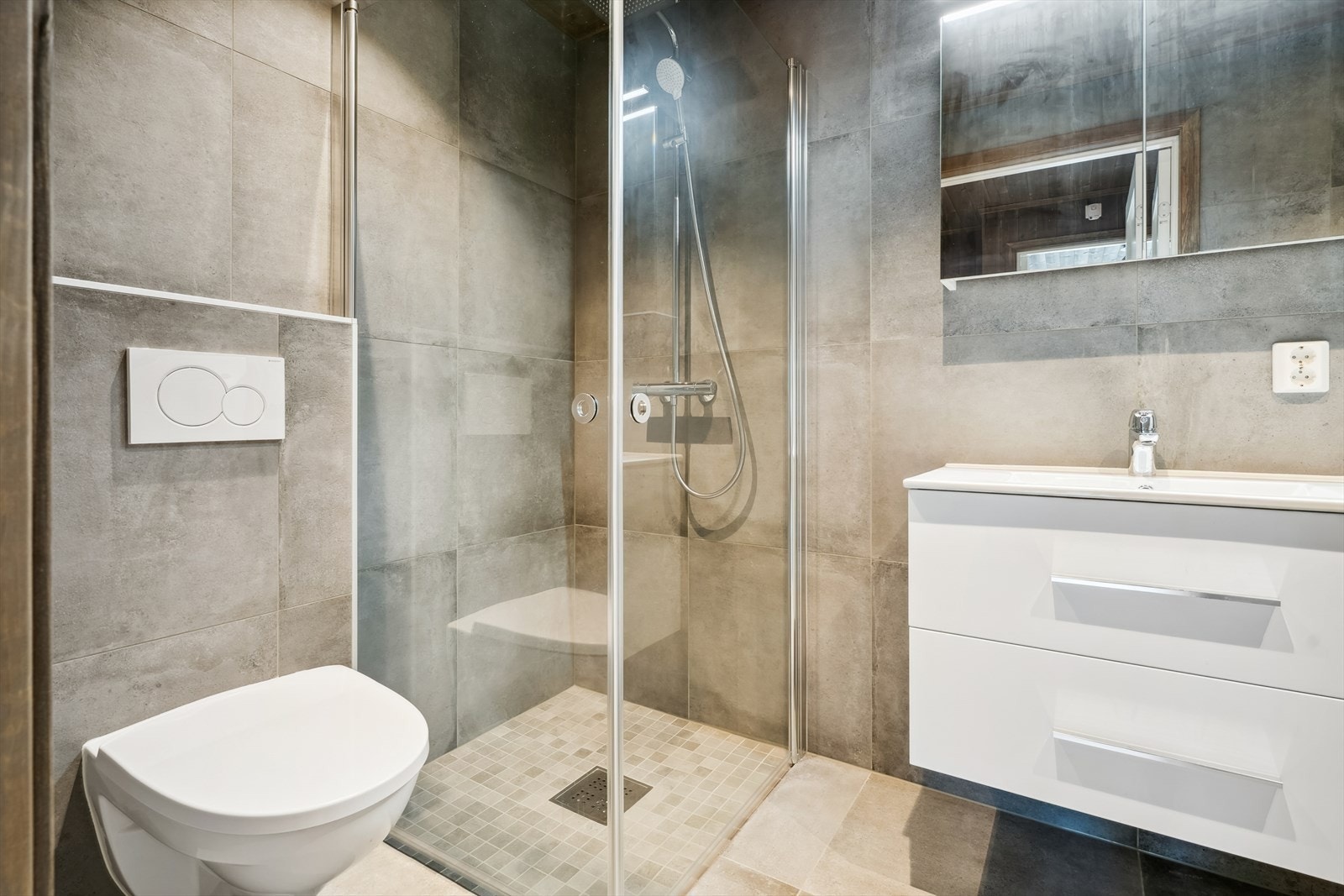 Pent flislagt baderom med servant i baderomsinnredning, vegghengt toalett og regnfallsdusj med innfellbare glassdører. Galleribilde