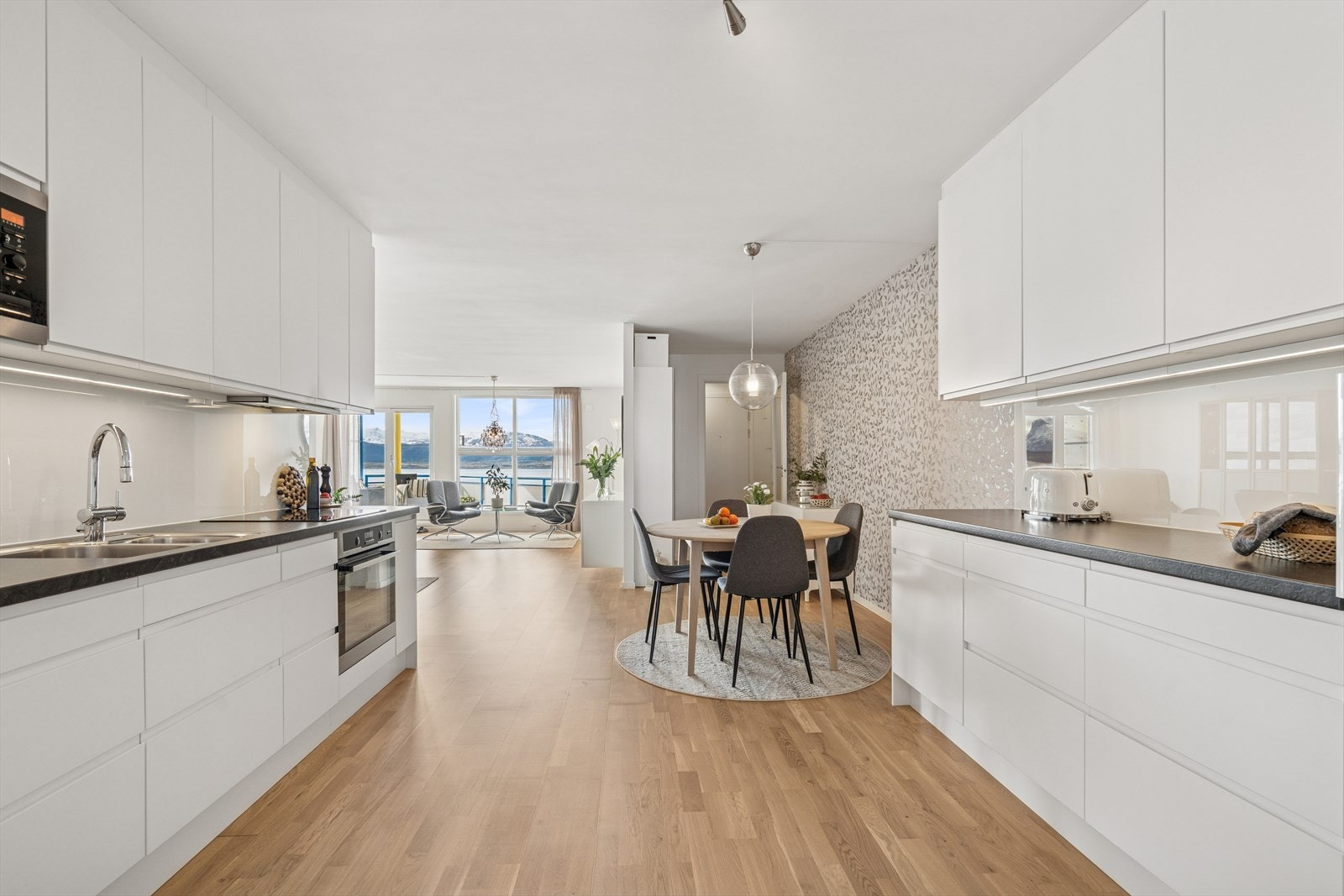 Kjøkkenet ligger i åpen løsning og har en moderne innredning bestående av hvite slette fronter m/grepslist, laminert benkeplate og glassplate på vegg over benk Galleribilde