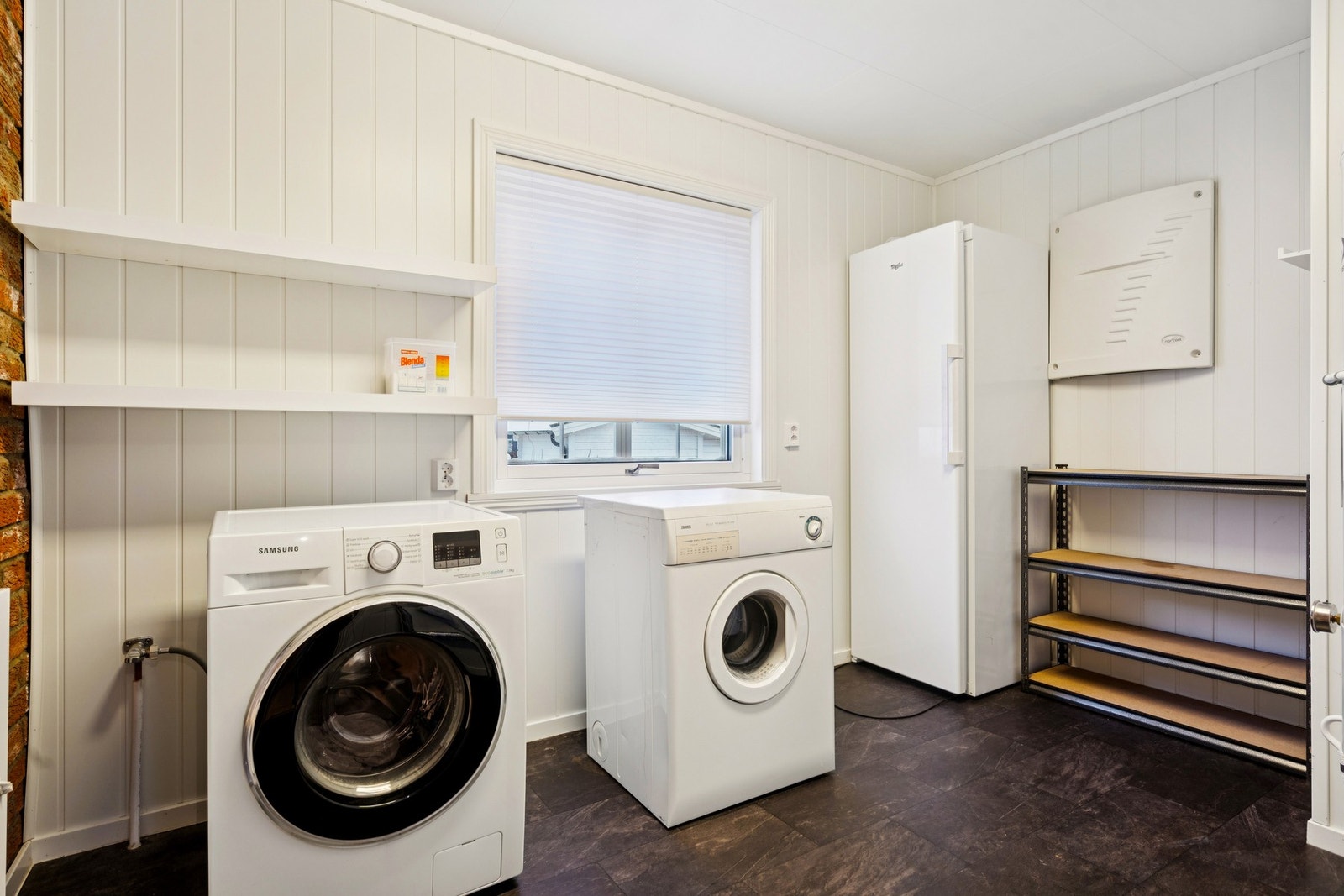 Veggene har MDF-panelplater og teglstein, taket har himlingsplater, gulvet er belagt med gulvbelegg uten oppkant, og rommet har både veggmonterte hyller og opplegg for vaskemaskin Galleribilde