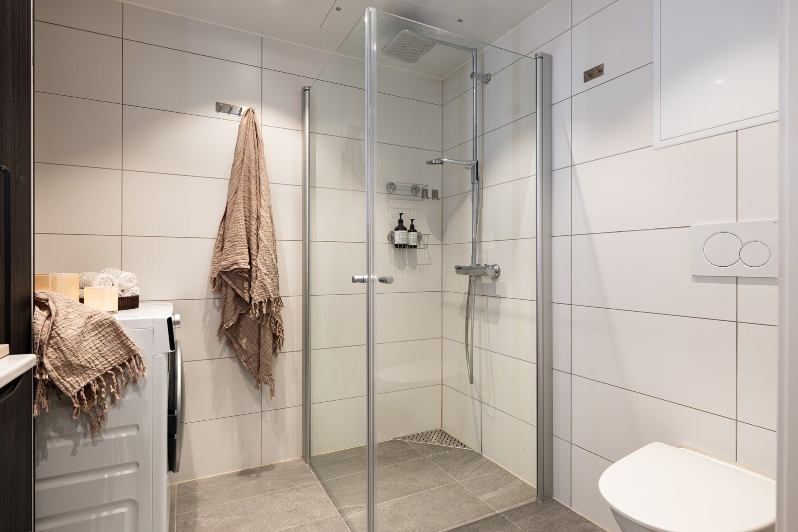 Baderommet er utstyrt med dusjdører av glass og dusjgarnityr på vegg, servant, opplegg for vaskemaskin og WC. Galleribilde