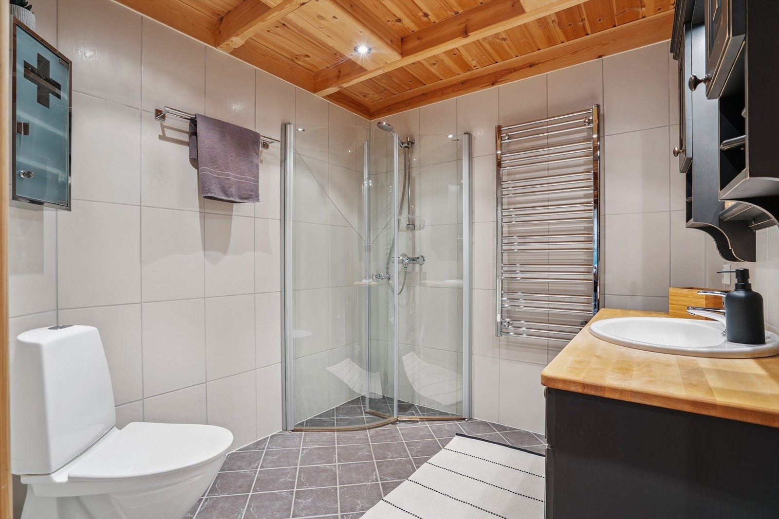 Flislagt baderom med elektriske varmekabler i gulv. Rommet inneholder gulvmontert toalett og dusjhjørne med innfellbare glassdører og veggmontert dusj. Galleribilde