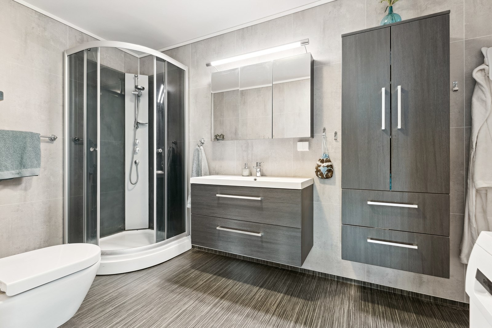 Stilreint bad innreiia med dusjkabinett, servant, vegghengt skap- og toalett, og eit stort speil med lys. Galleribilde